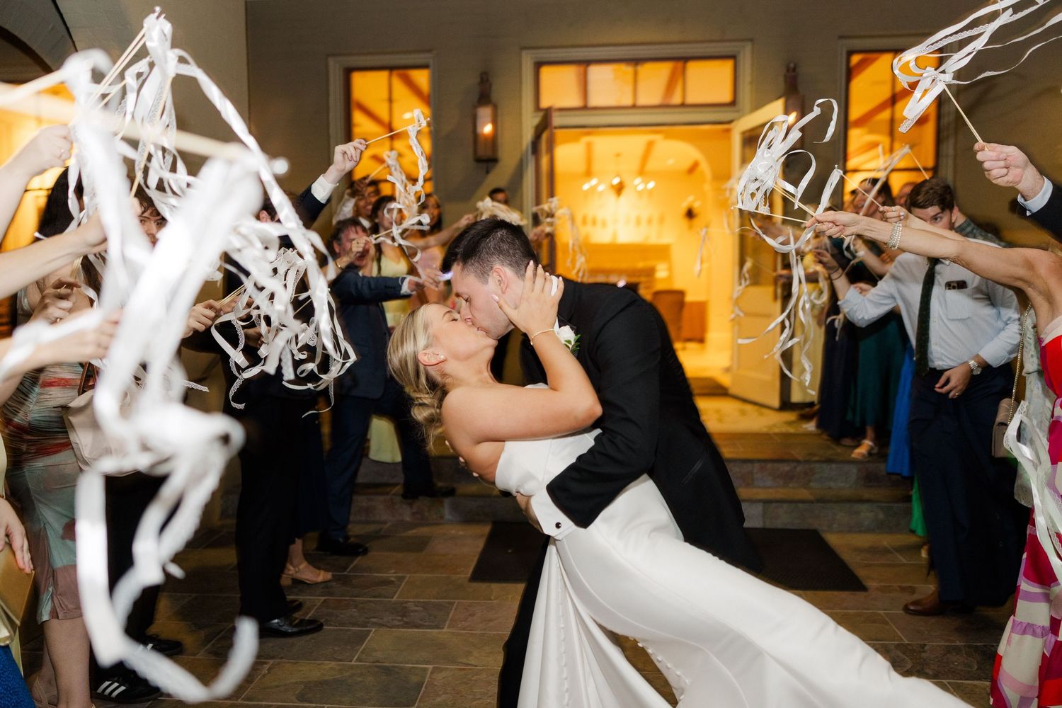 Wedding exit celebration with guests waving white streamers in evening lighting.
