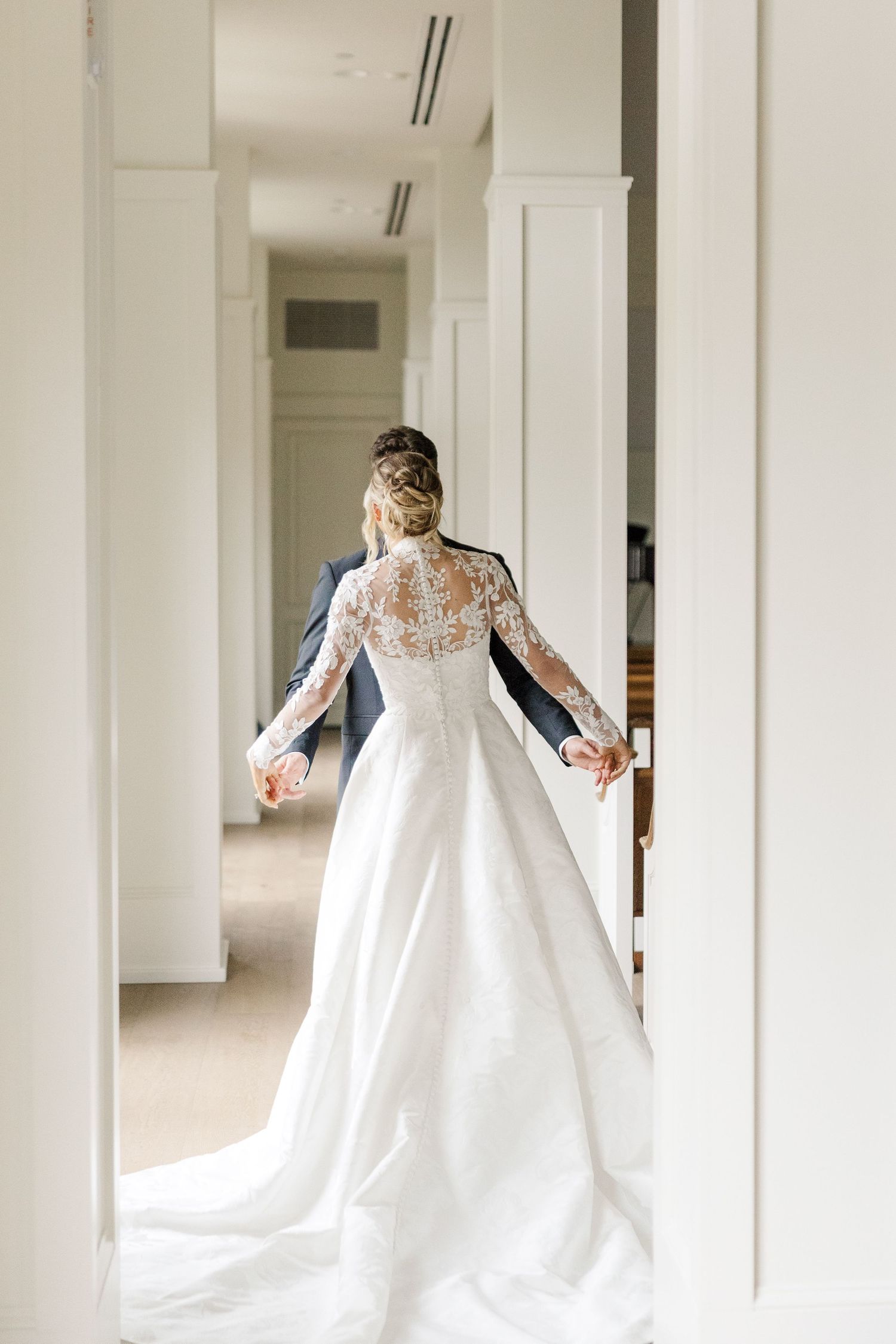 Elegant bride in flowing lace wedding gown walking through bright white hallway.