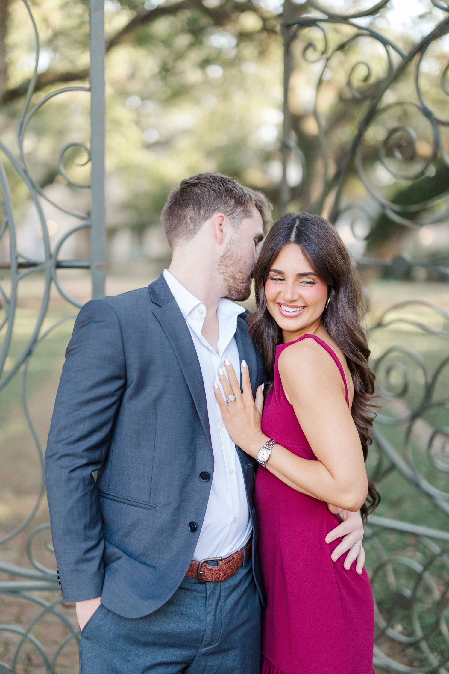 A romantic couple embraces by an ornate iron gate, with her wearing a burgundy dress and him in a gray suit.