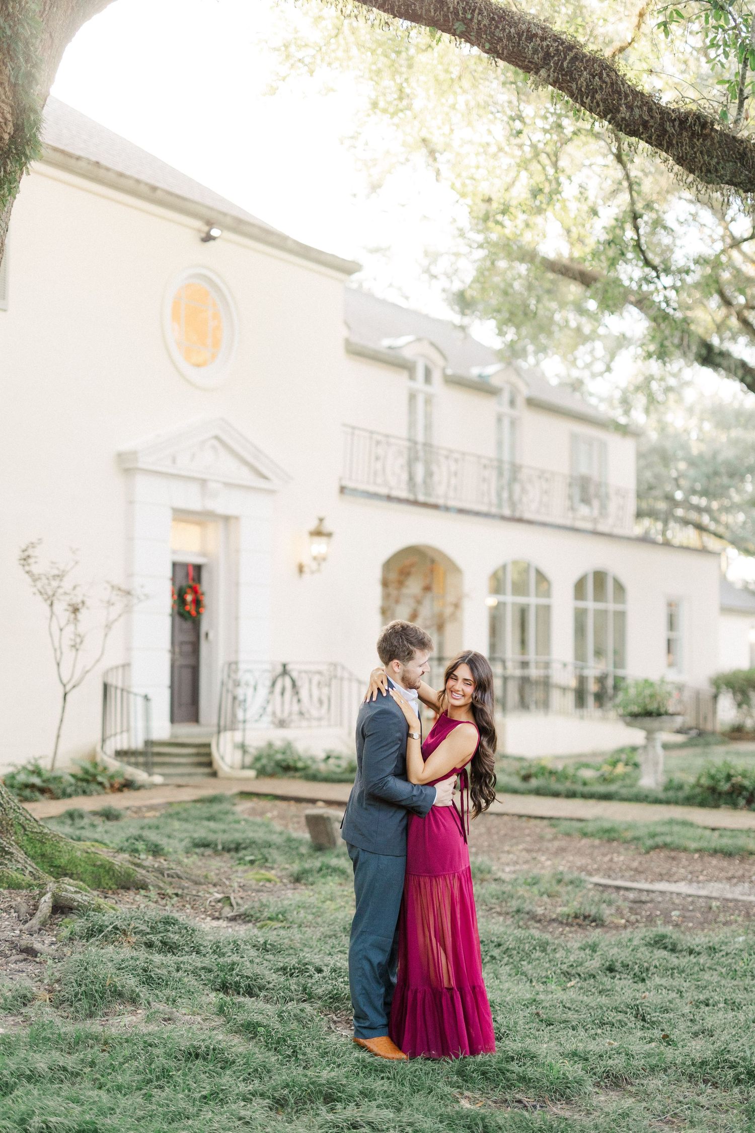 Elegant couple embracing on landscaped grounds with a white building in the background.