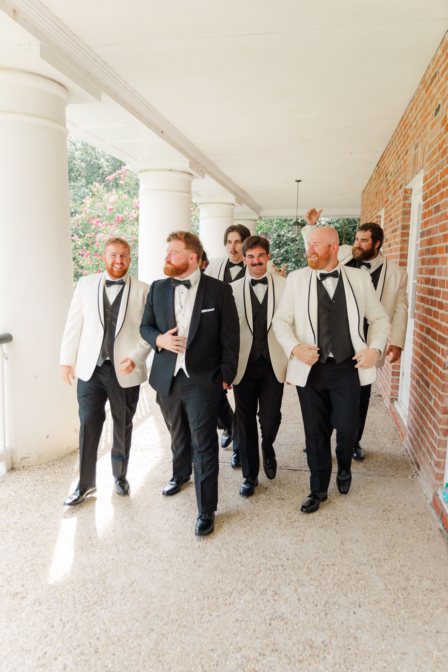 Groomsmen in black and white formal attire walking together through a columned hallway.