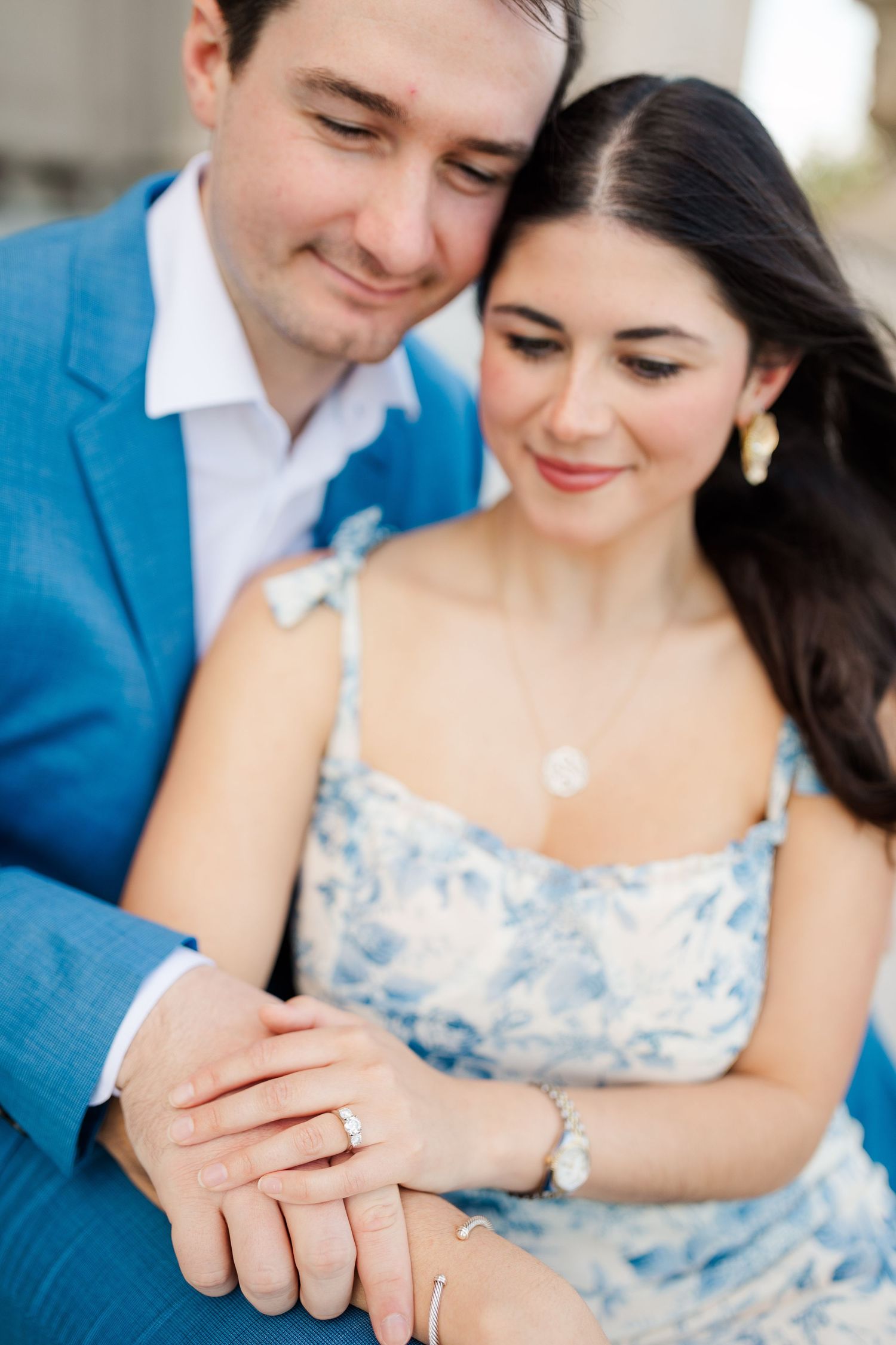 A couple in formal attire share a tender moment showing their wedding rings against a blue floral dress and suit.