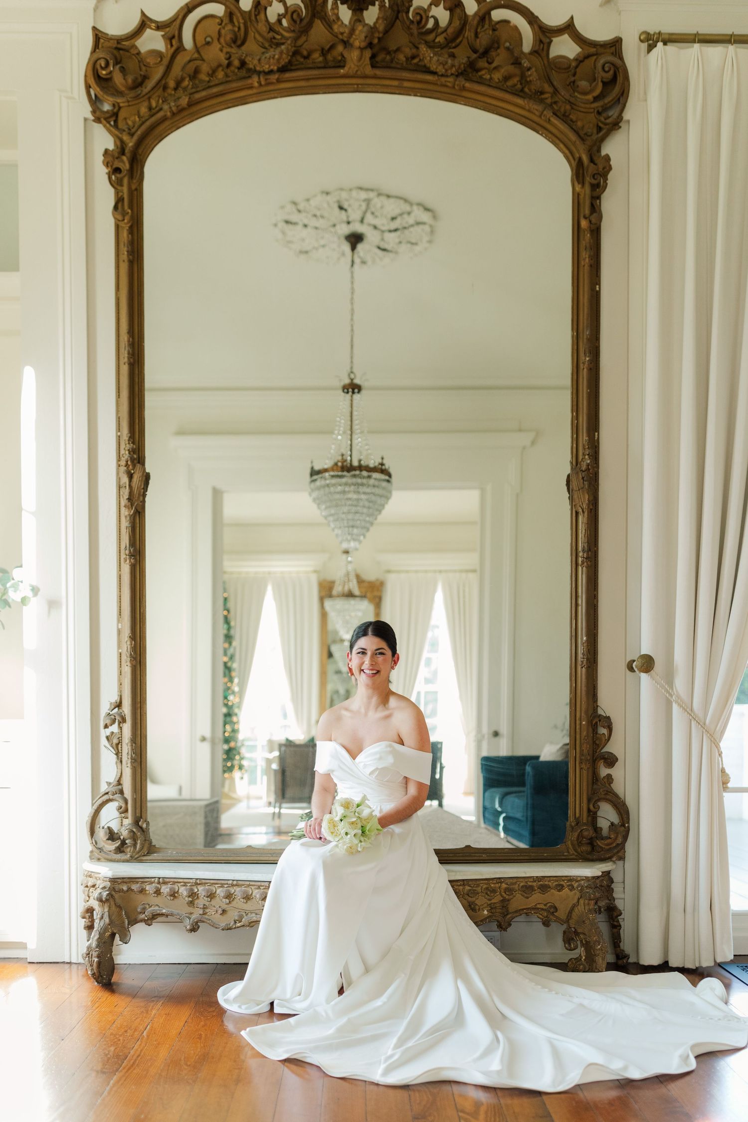 A bride stands gracefully before an ornate gold mirror in a classic off-shoulder wedding gown with a flowing train.