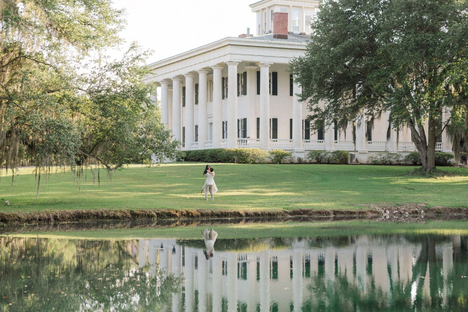 Couple poses by a reflective pond in front of a grand white plantation mansion with tall columns.