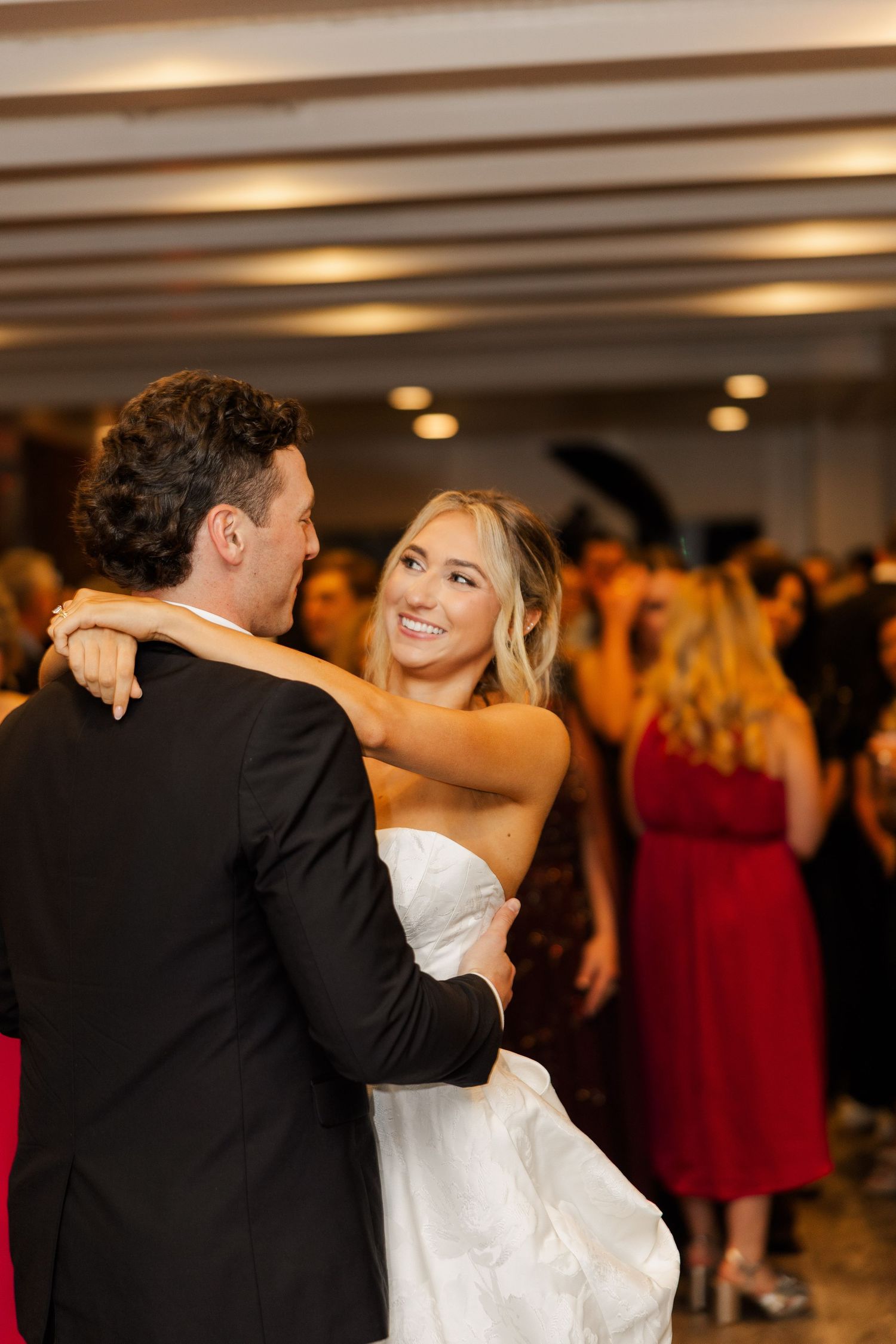 Newlyweds dance together at their reception surrounded by guests in formal attire.