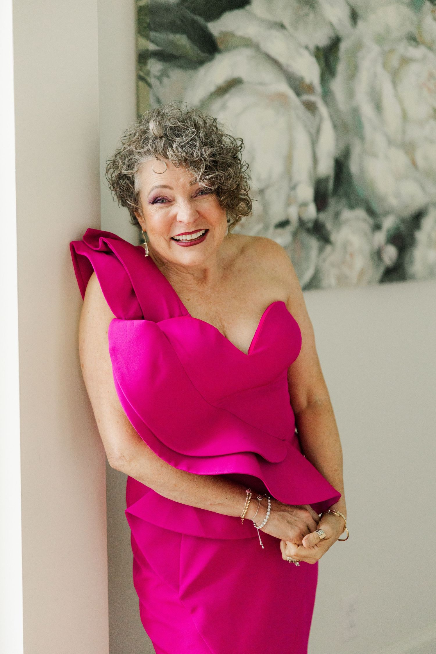 Woman wearing vibrant hot pink one-shoulder dress smiles joyfully in front of abstract art.