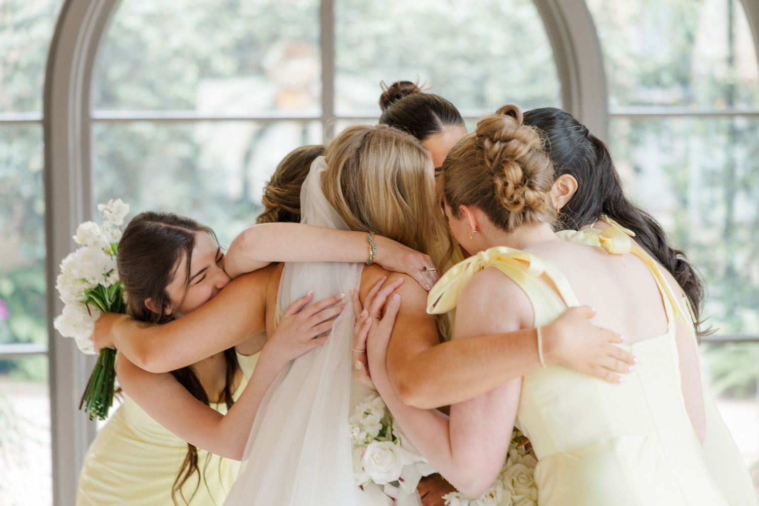 A row of bridesmaids in light yellow dresses embrace in a group hug during a wedding celebration.