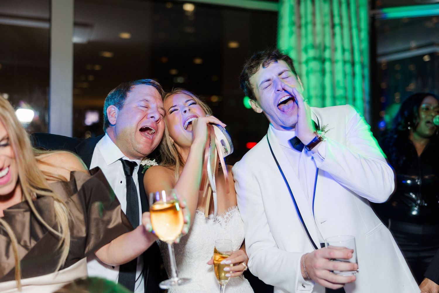 Wedding guests laughing and celebrating with drinks at a reception with green ambient lighting.