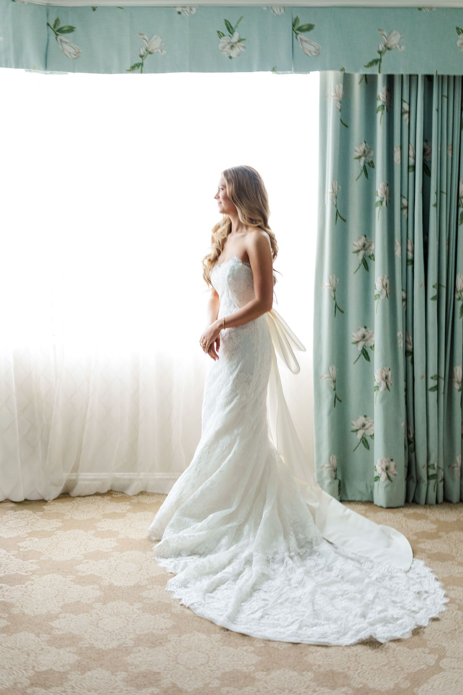 A bride in a flowing white lace wedding gown poses elegantly against turquoise floral curtains in a sunlit room.