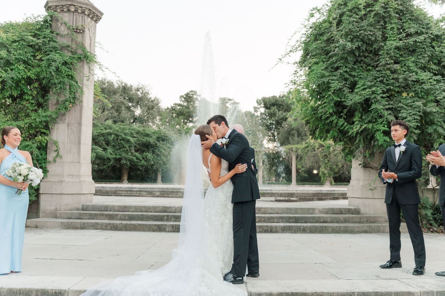 Wedding party takes formal photos on stone steps in an outdoor garden setting.