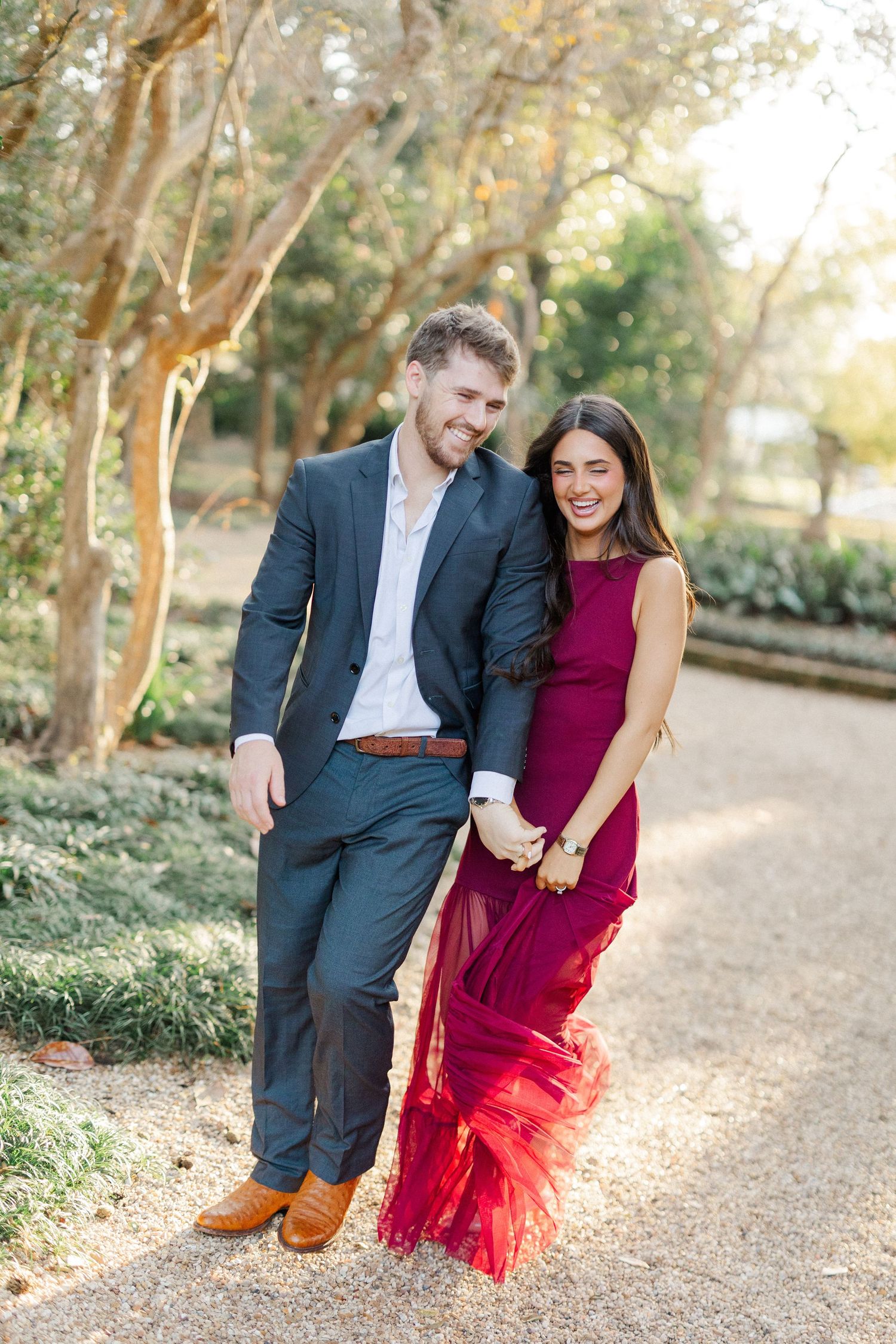 A couple in formal attire walks together on a sunlit path through trees during an autumn engagement photo session.
