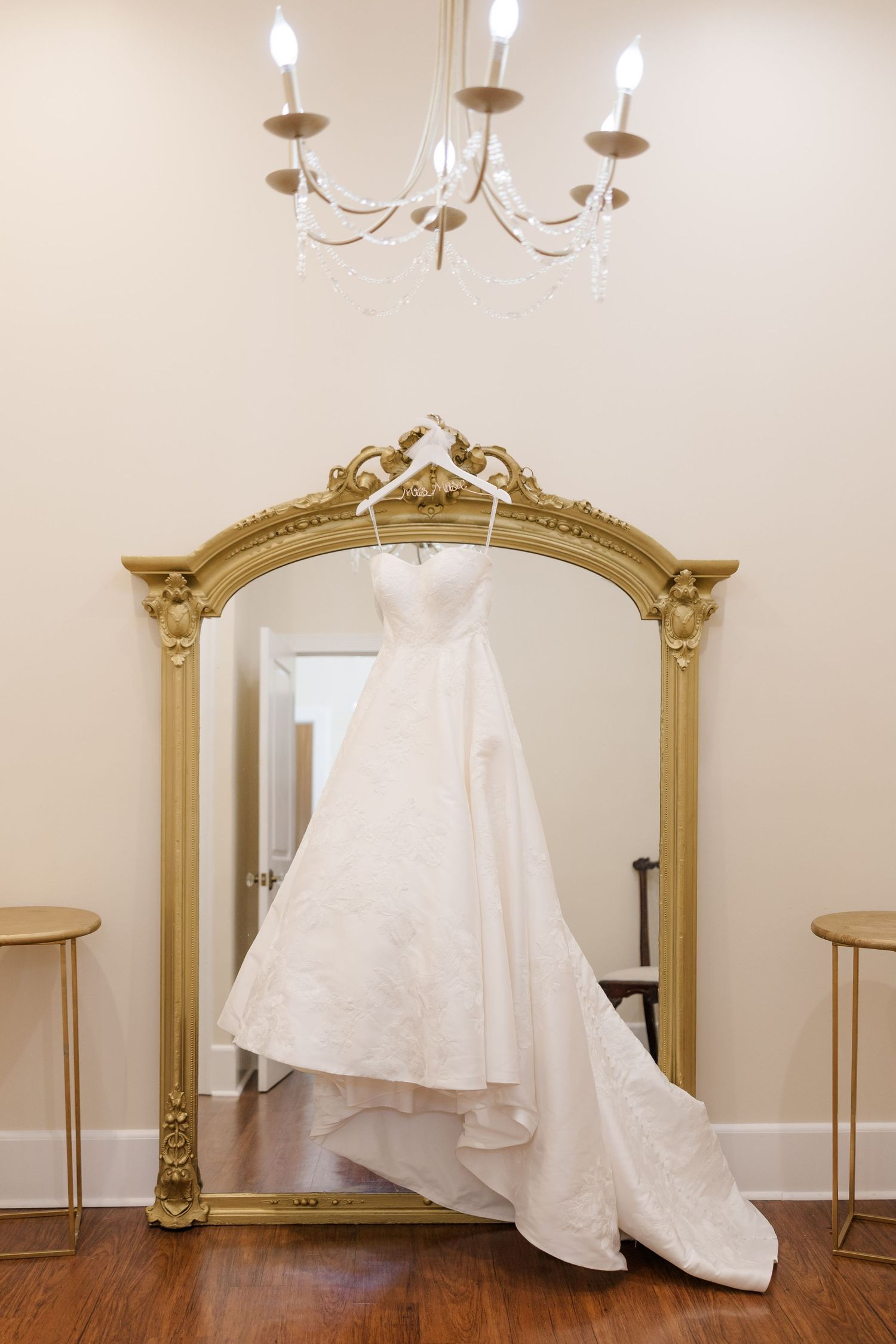 A white wedding dress hangs elegantly against a beige wall with ornate gold-framed mirrors.