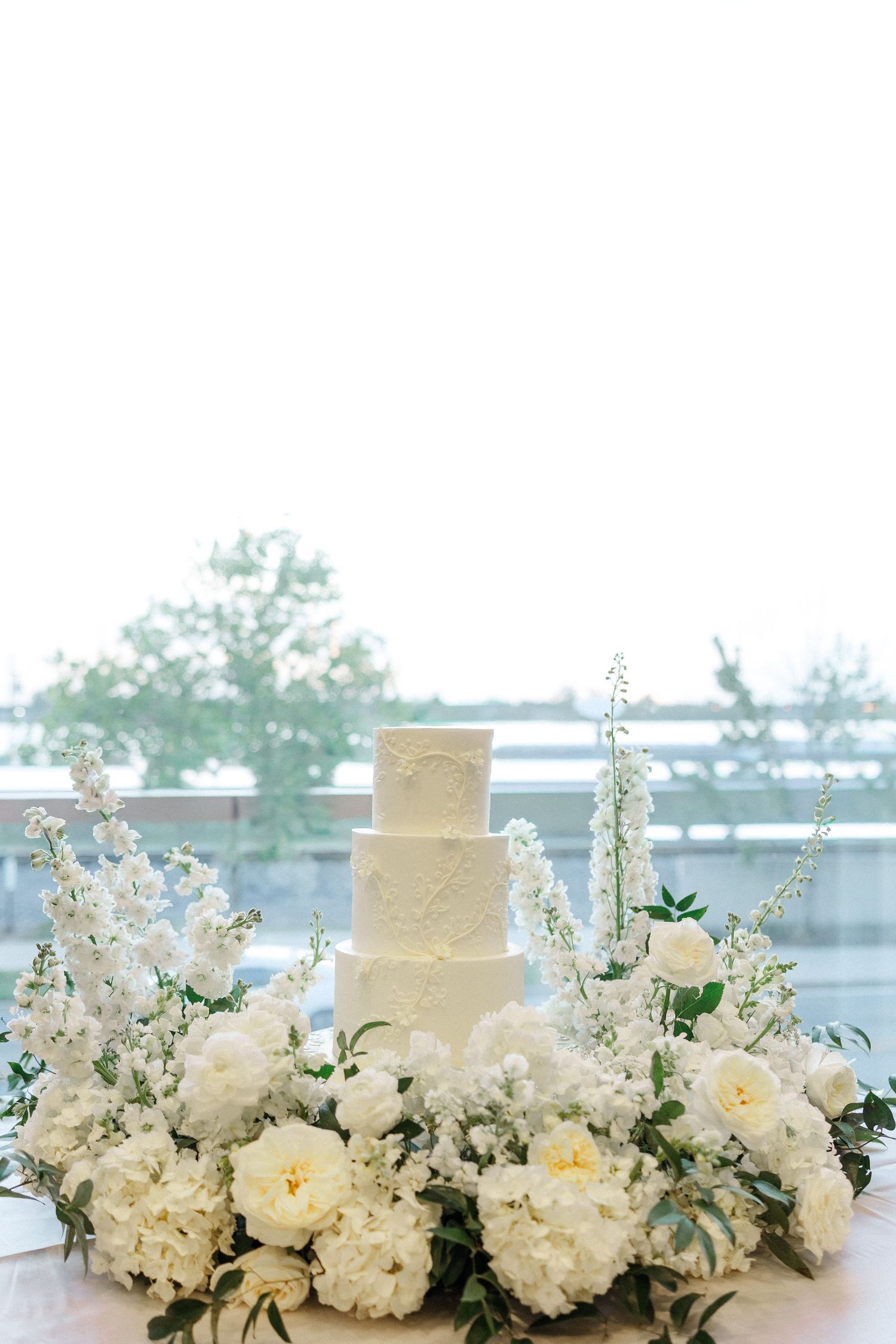 Four-tier white wedding cake surrounded by lush white peonies and florals on a table with outdoor views.