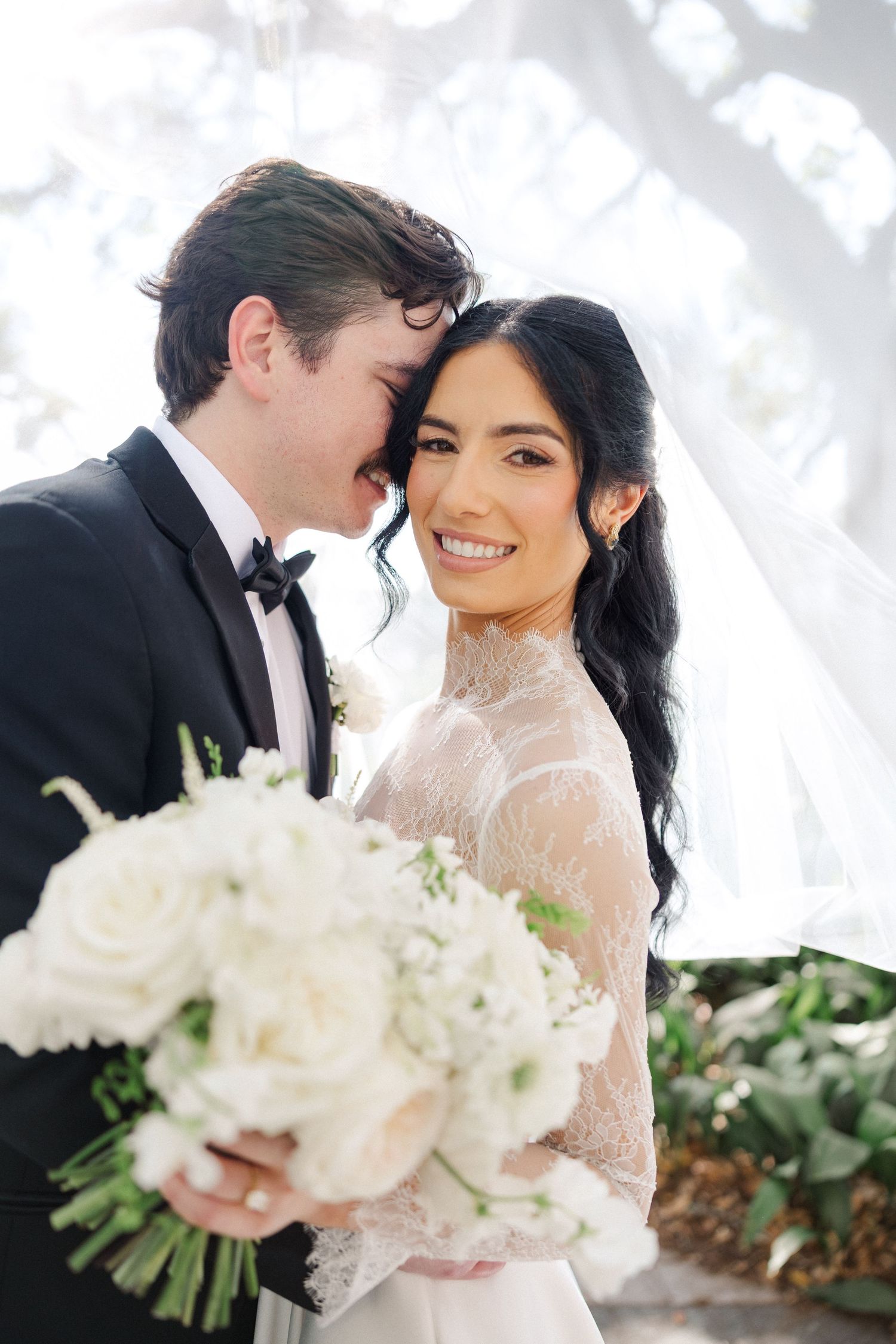 Groom whispers to smiling bride holding white rose bouquet on their wedding day.