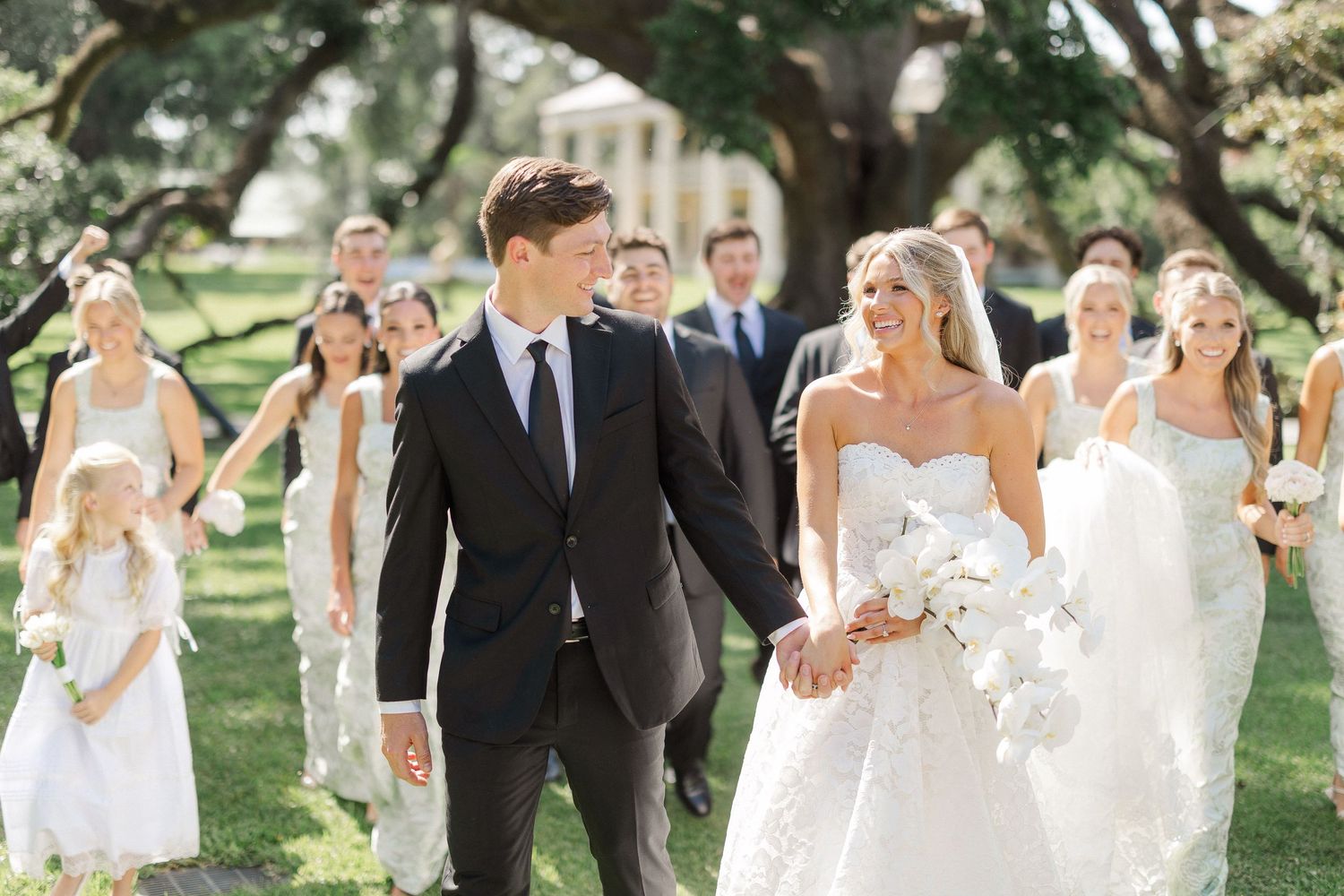 Bride and groom walk hand-in-hand with wedding party in white dresses and black suits on a sunny outdoor lawn.