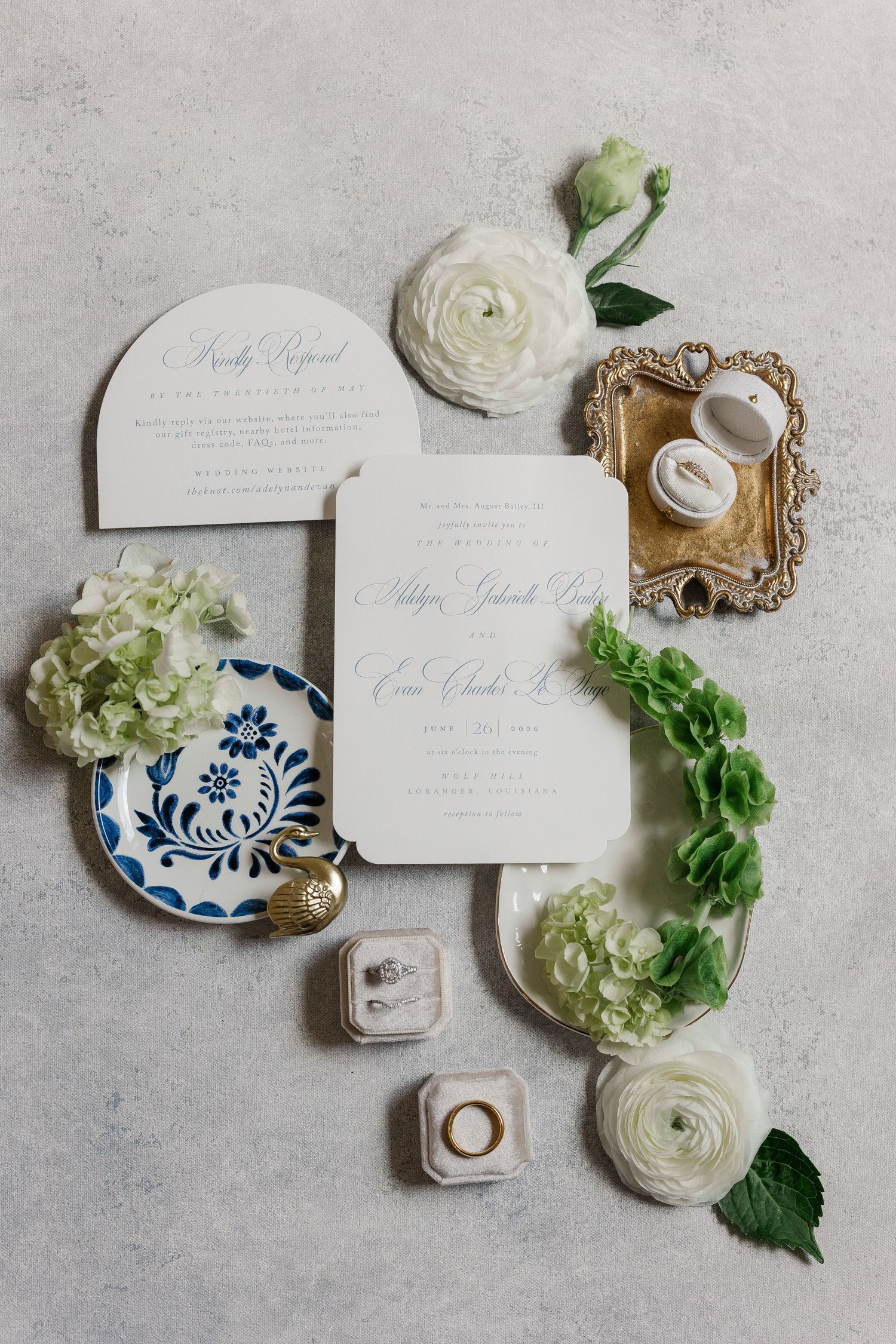 Elegant wedding invitation suite with white florals, blue ceramic dish, gold ring box, and rings on gray background.