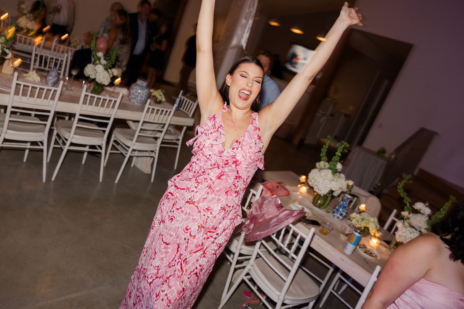 Joyful wedding guest in pink floral dress celebrates with arms raised on the reception dance floor.