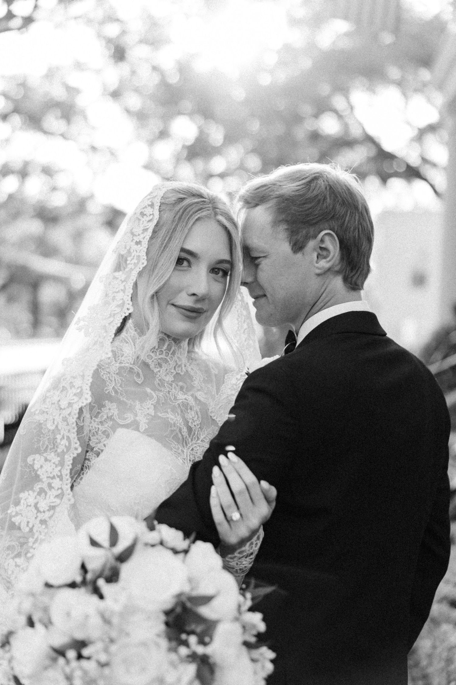 Black and white photo of a bride in lace veil and groom in tuxedo embracing, holding a floral bouquet.