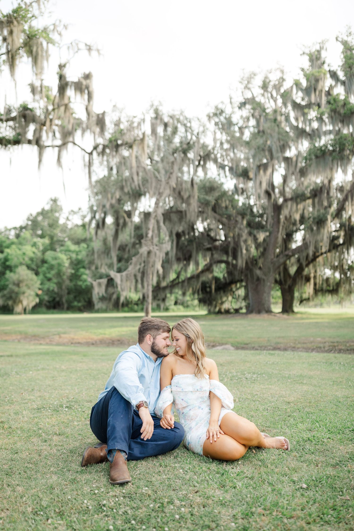 A couple sits together on grass beneath large oak trees draped with Spanish moss in a romantic southern garden setting.