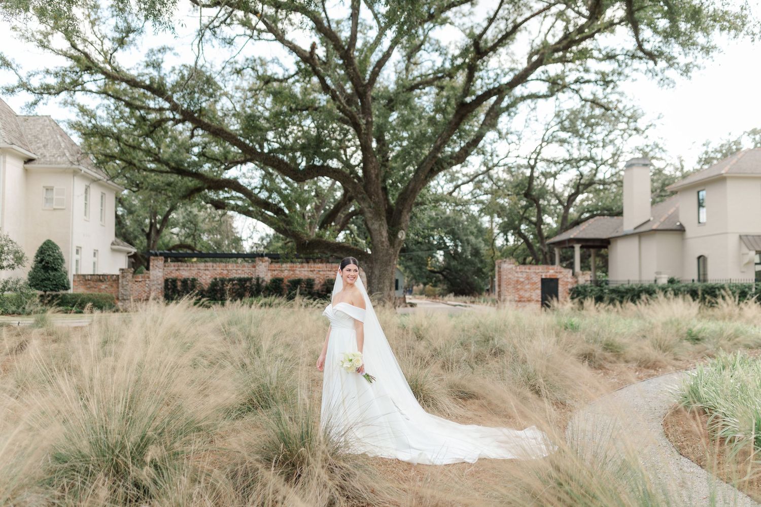 A flowy wedding gown creates an elegant silhouette against tall grass and a majestic oak tree at an outdoor venue.