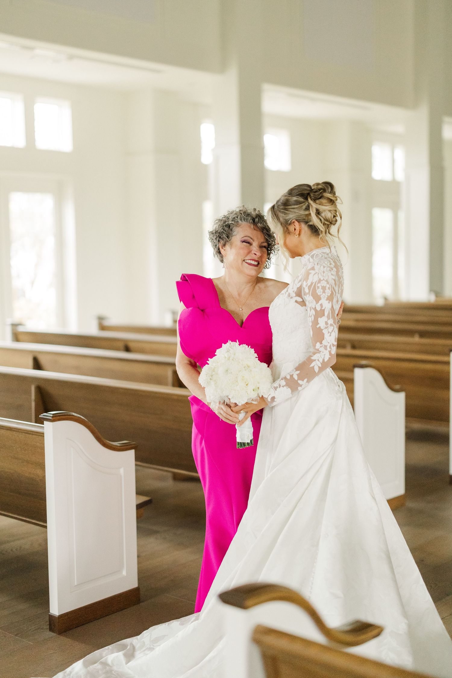 Emotional moment between bride in lace gown and guest in bright pink dress in church setting.