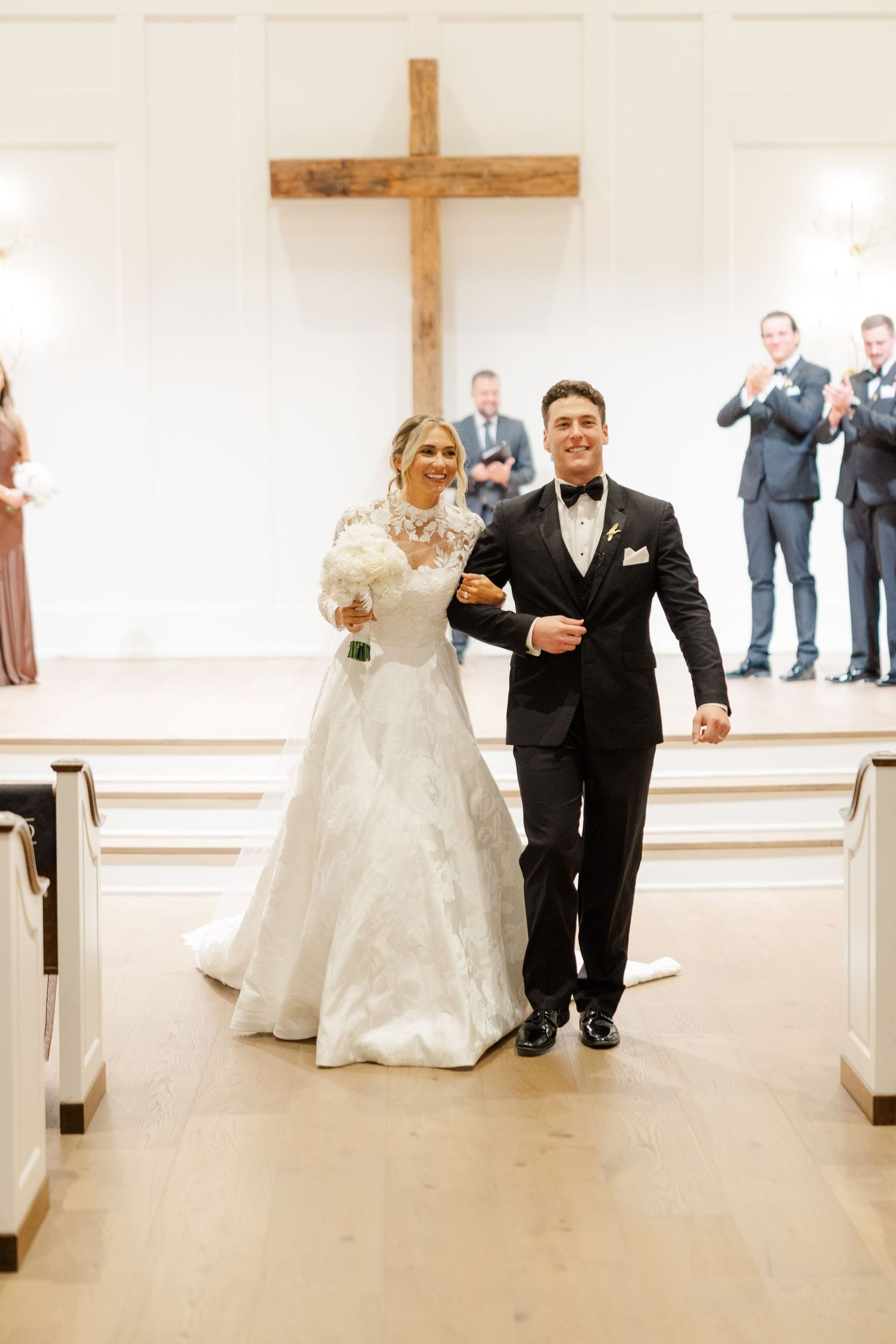 A couple walks down the church aisle after their wedding ceremony with a cross visible in the background.