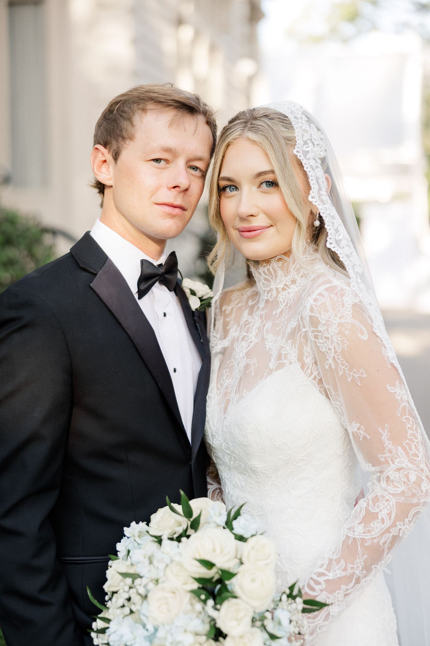 Classic wedding portrait featuring elegant lace gown and white floral bouquet against white architecture.