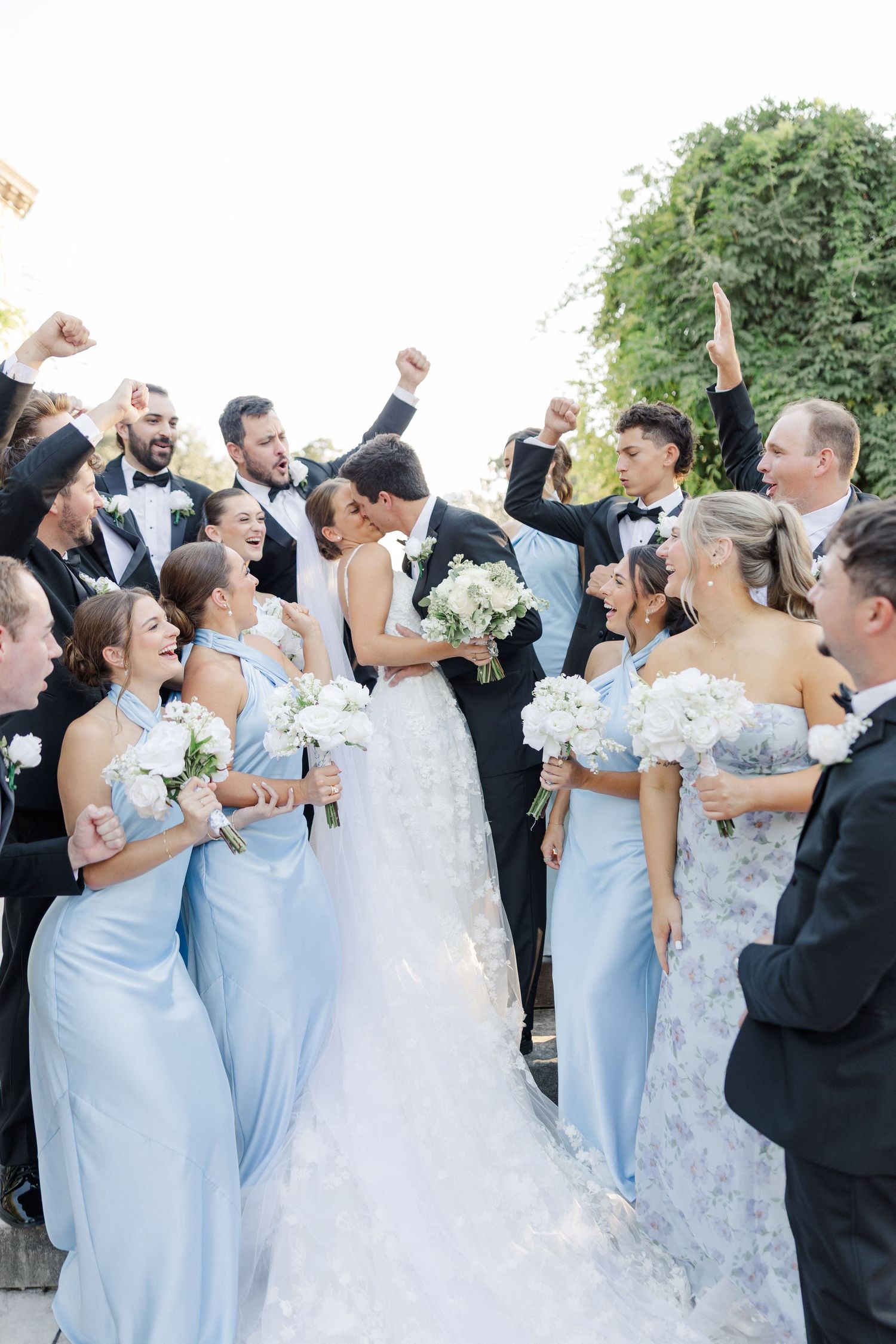 Wedding party celebrates joyfully with bridesmaids in light blue dresses and white bouquets outdoors.
