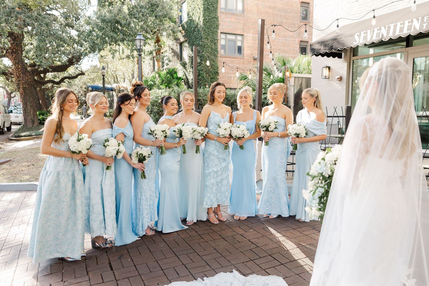 Bridesmaids in light blue dresses pose together with the bride in a sunny outdoor setting.