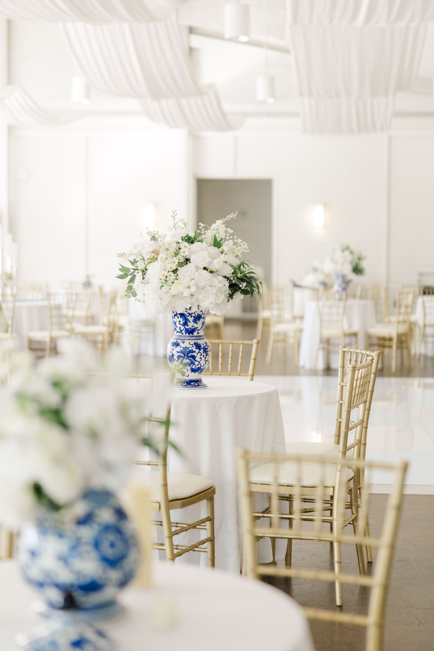 Elegant white wedding reception venue with gold chiavari chairs and blue and white floral centerpieces.