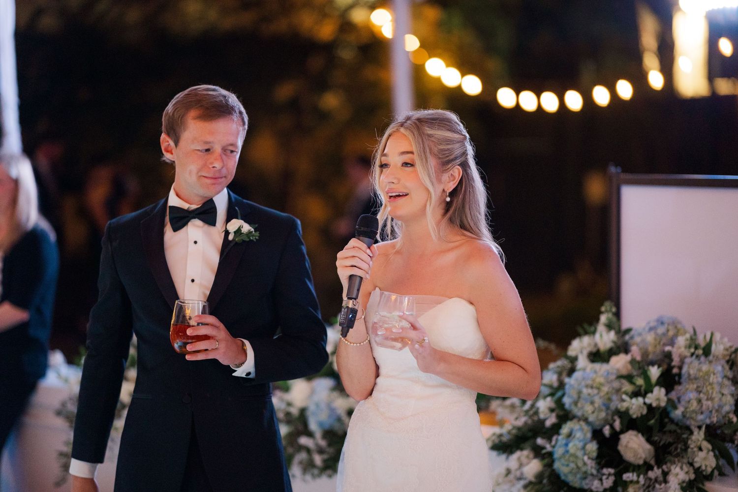 Wedding couple giving speeches during their evening reception with string lights in background.