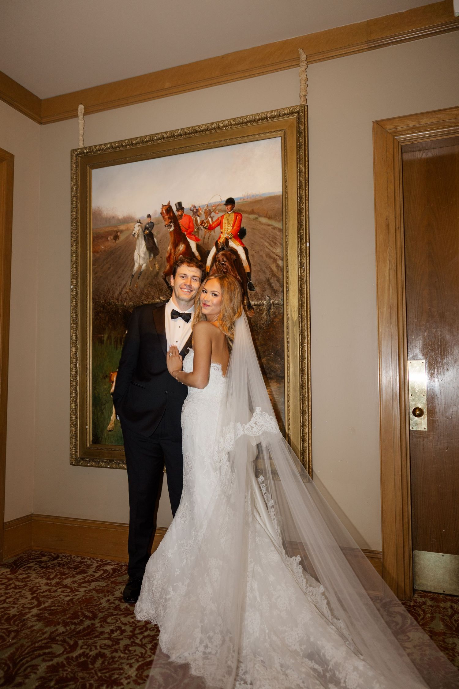 A couple in wedding attire share playful moments in a hallway with framed artwork.