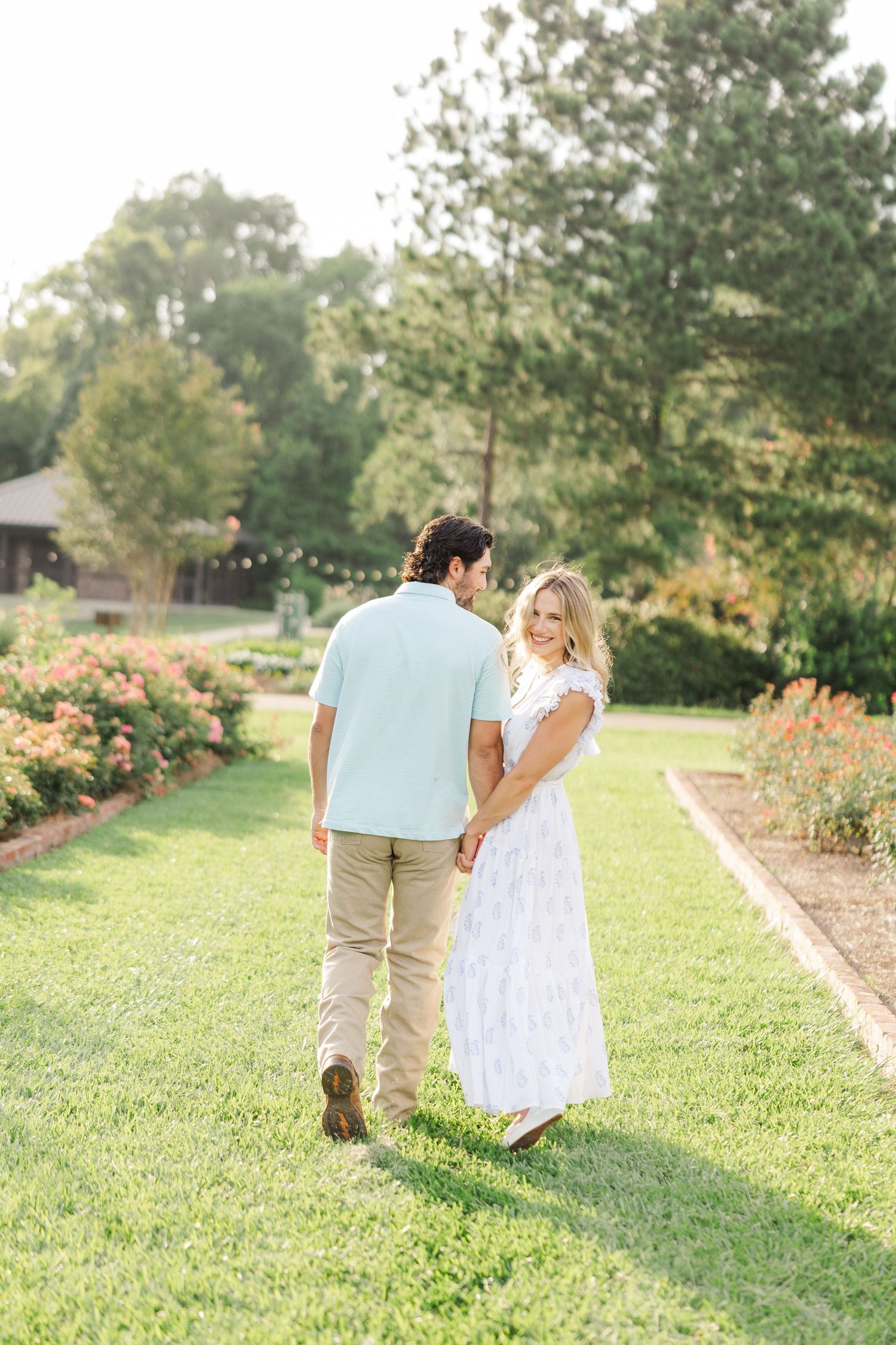 A couple strolls hand in hand through a garden pathway with green grass and trees in the background.