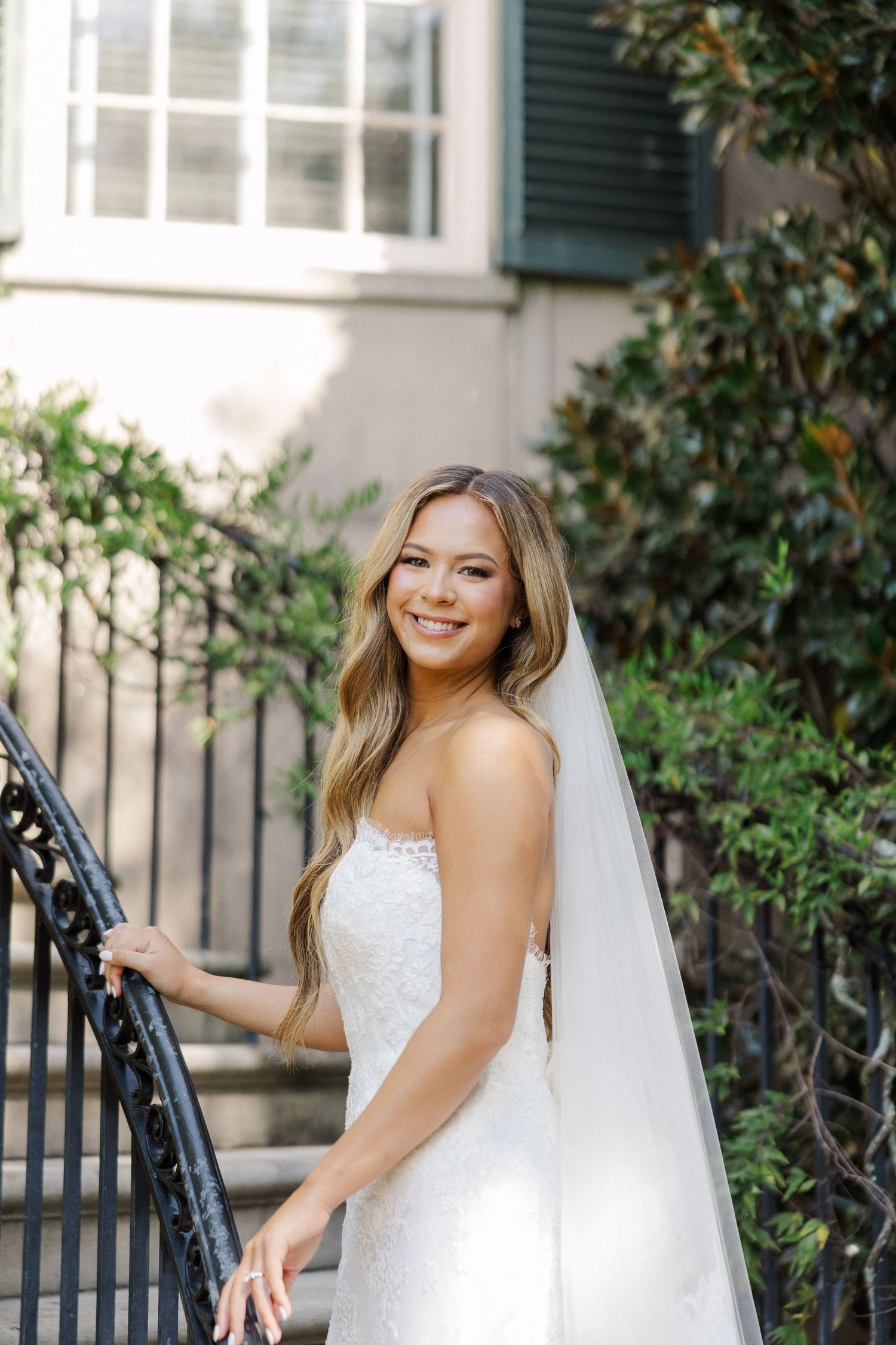 A bride in a white lace wedding dress poses elegantly against a white house with green shutters and climbing vines.