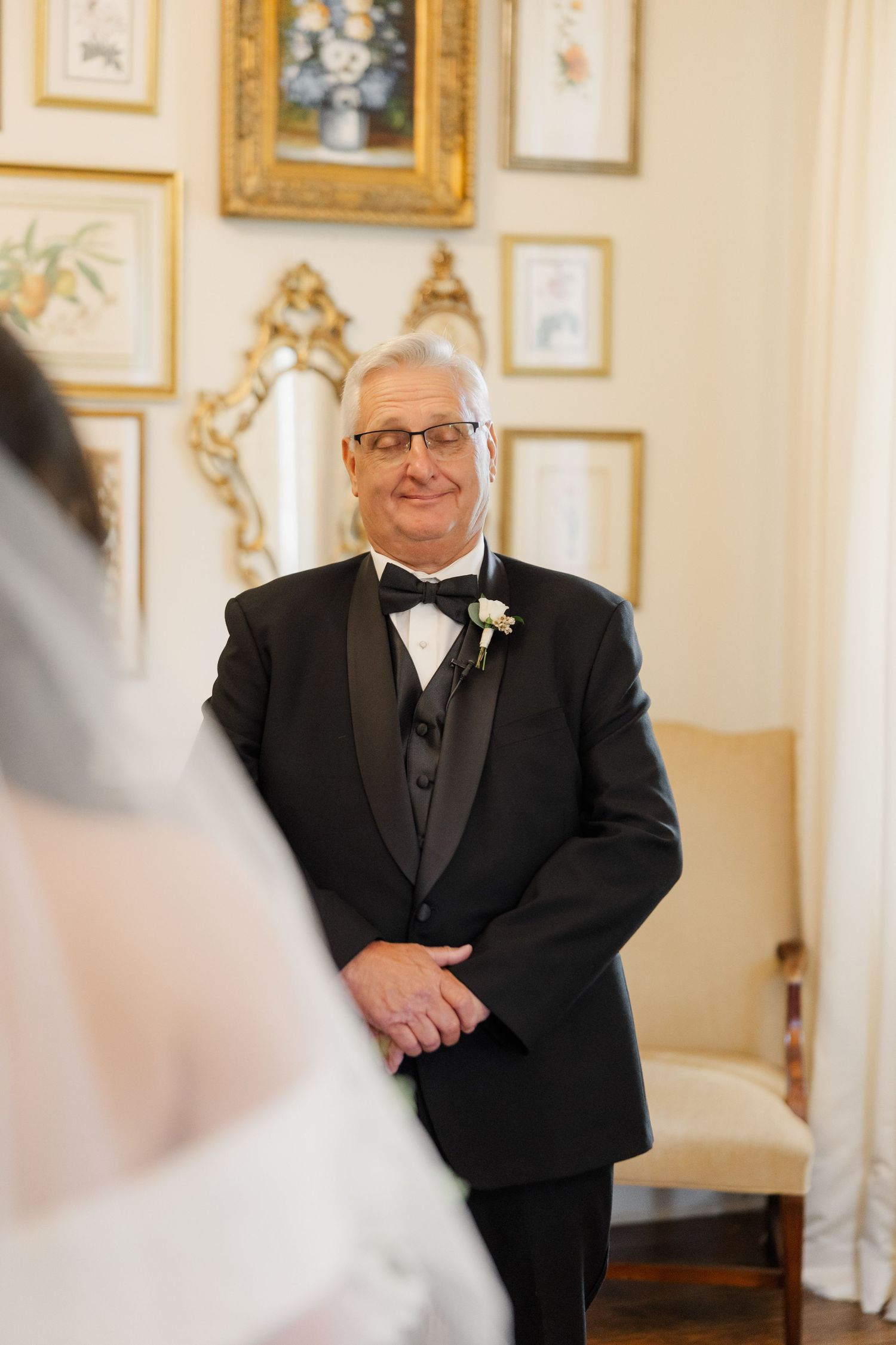 A person wearing a black tuxedo with bow tie and glasses smiles warmly in an elegant room with framed artwork on walls.