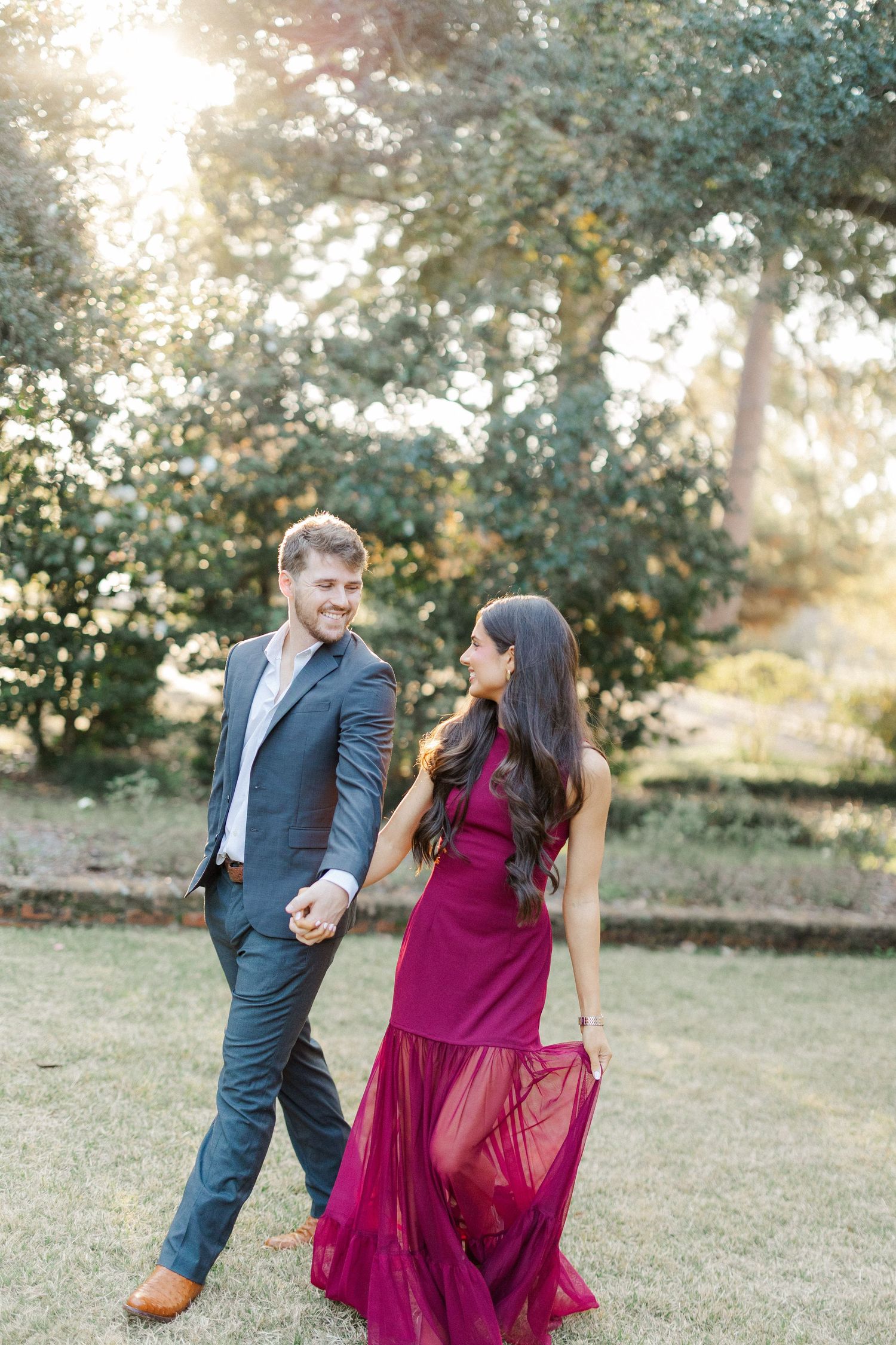 A couple walks together in a garden setting at sunset, with her wearing a flowing burgundy dress and him in a navy suit.