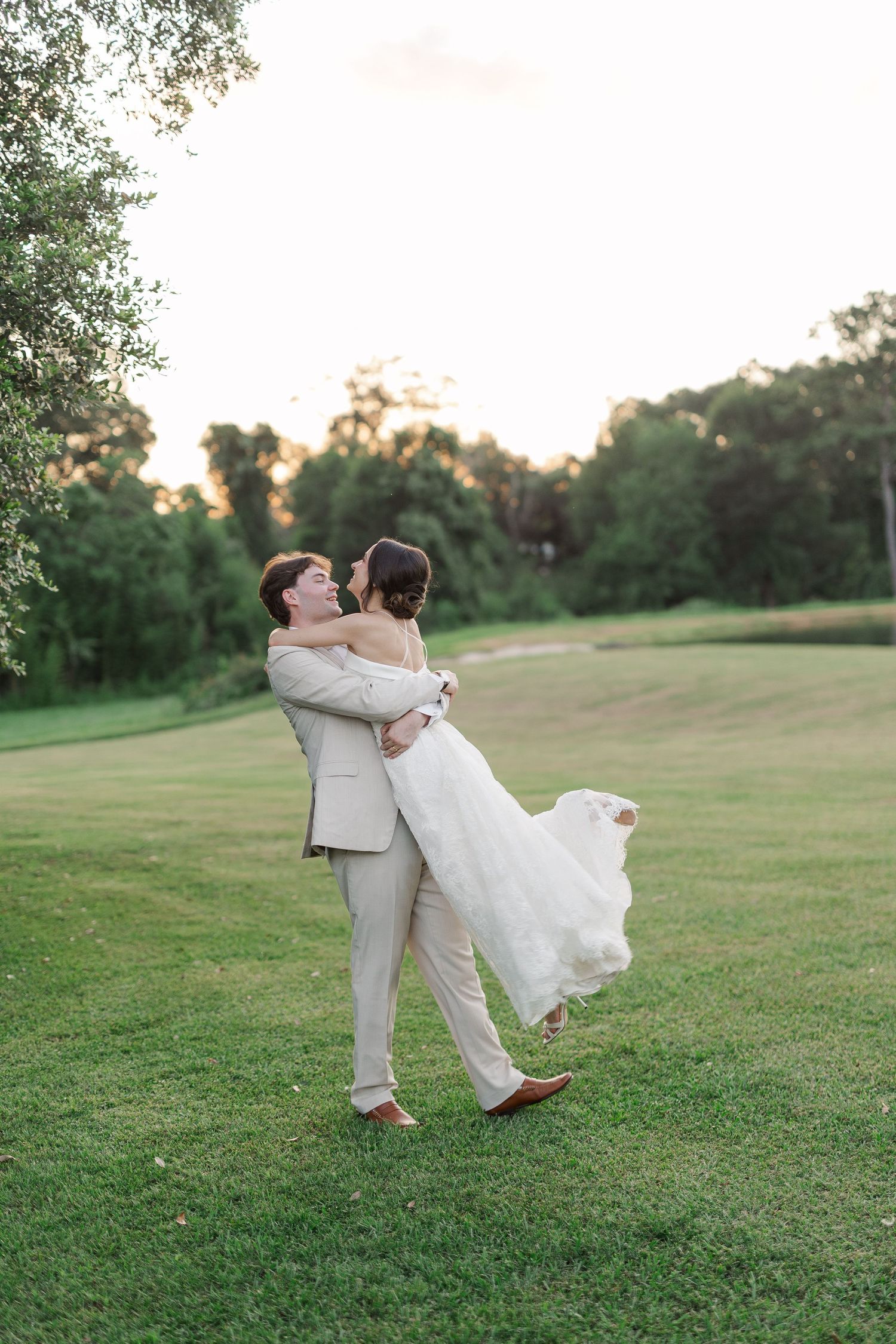 Joyful groom lifting bride in white lace gown at golden hour on lush green lawn.