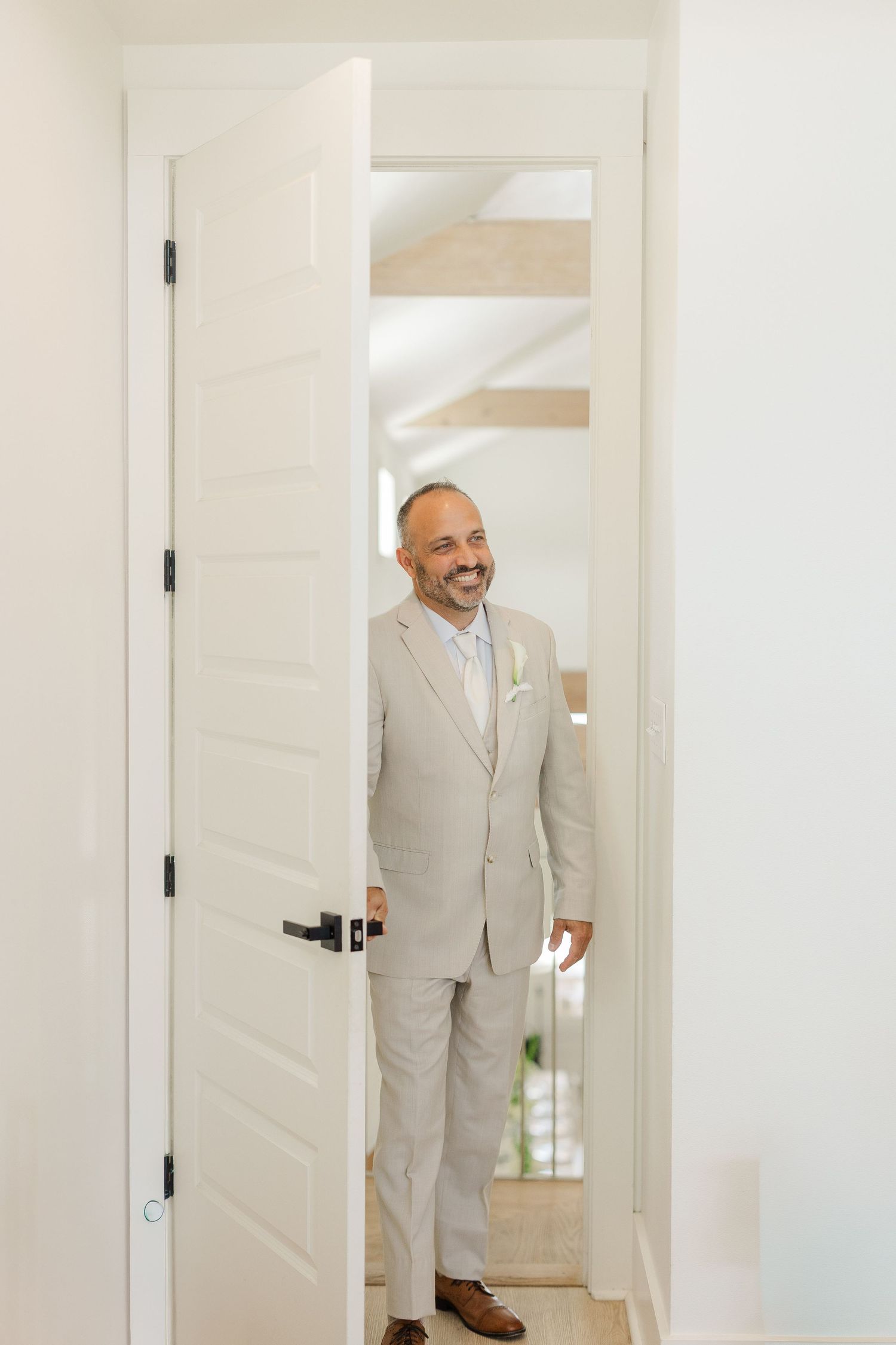 Smiling groom in beige suit with boutonniere standing in doorway during wedding day preparations.