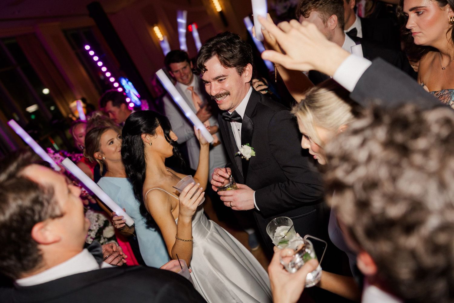 Bride and groom dancing joyfully with guests holding glow sticks at lively wedding reception.