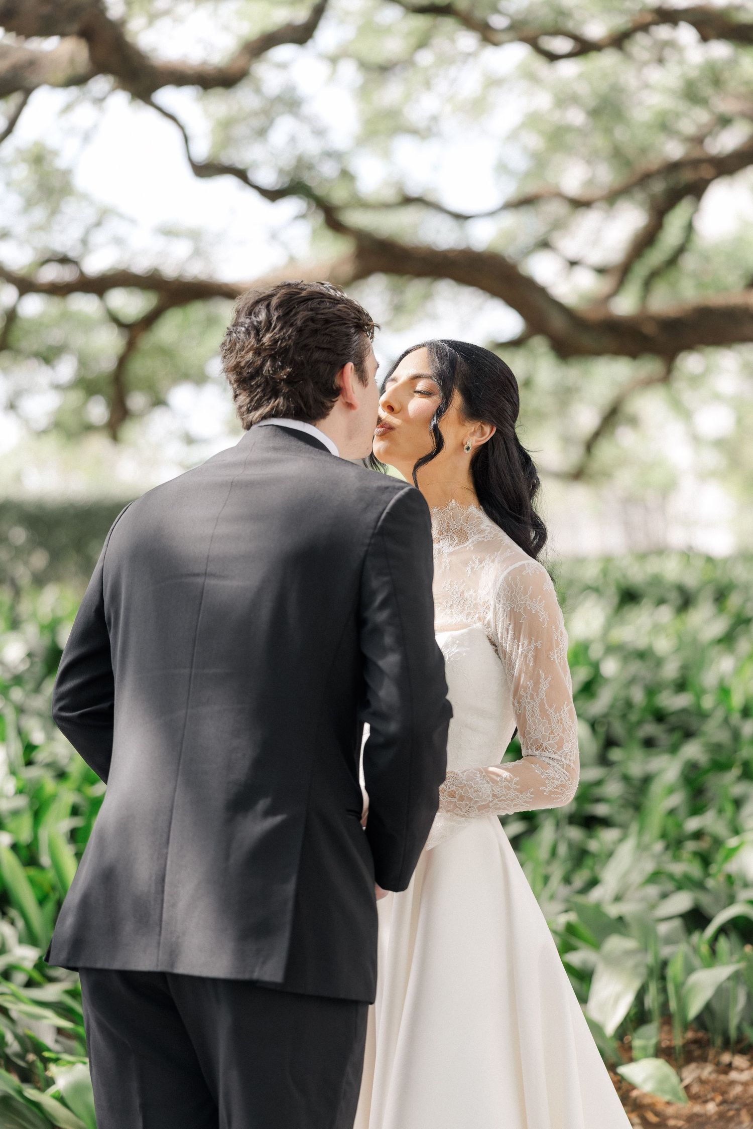 Bride in lace long-sleeve wedding gown kissing groom in black suit under sprawling oak tree.