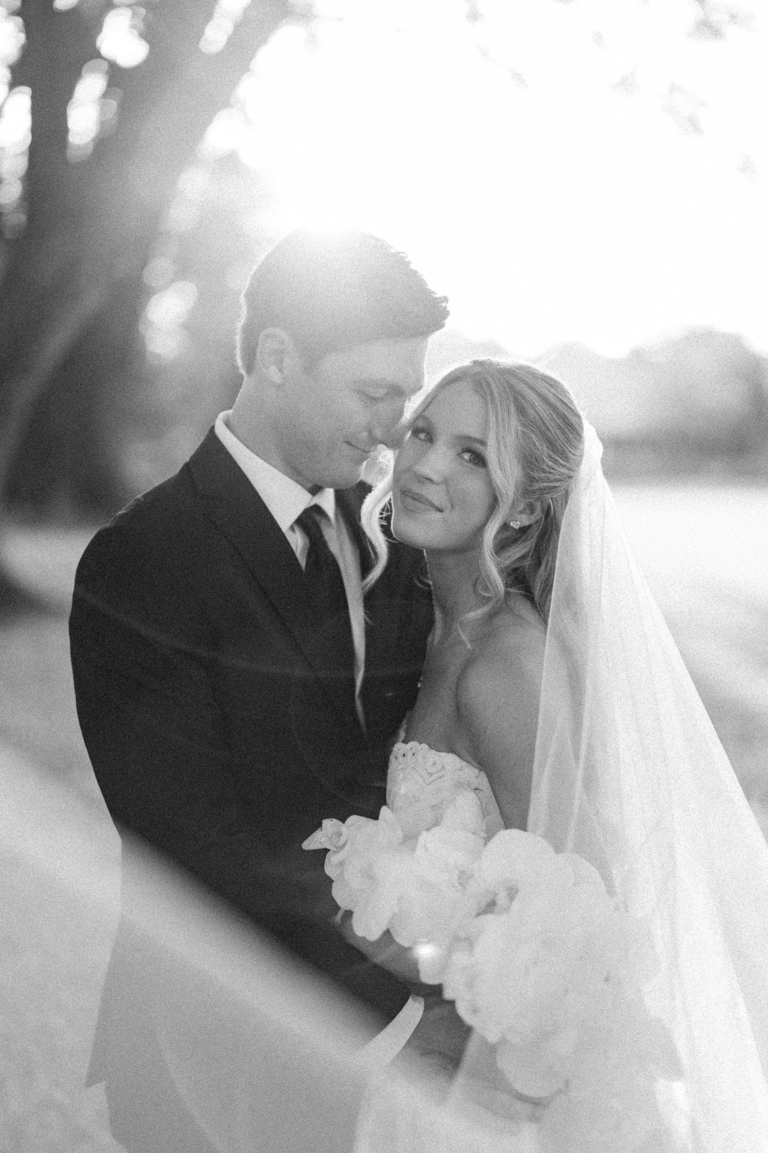 Black and white romantic portrait of bride and groom embracing at golden hour with soft sunlight glowing behind them.