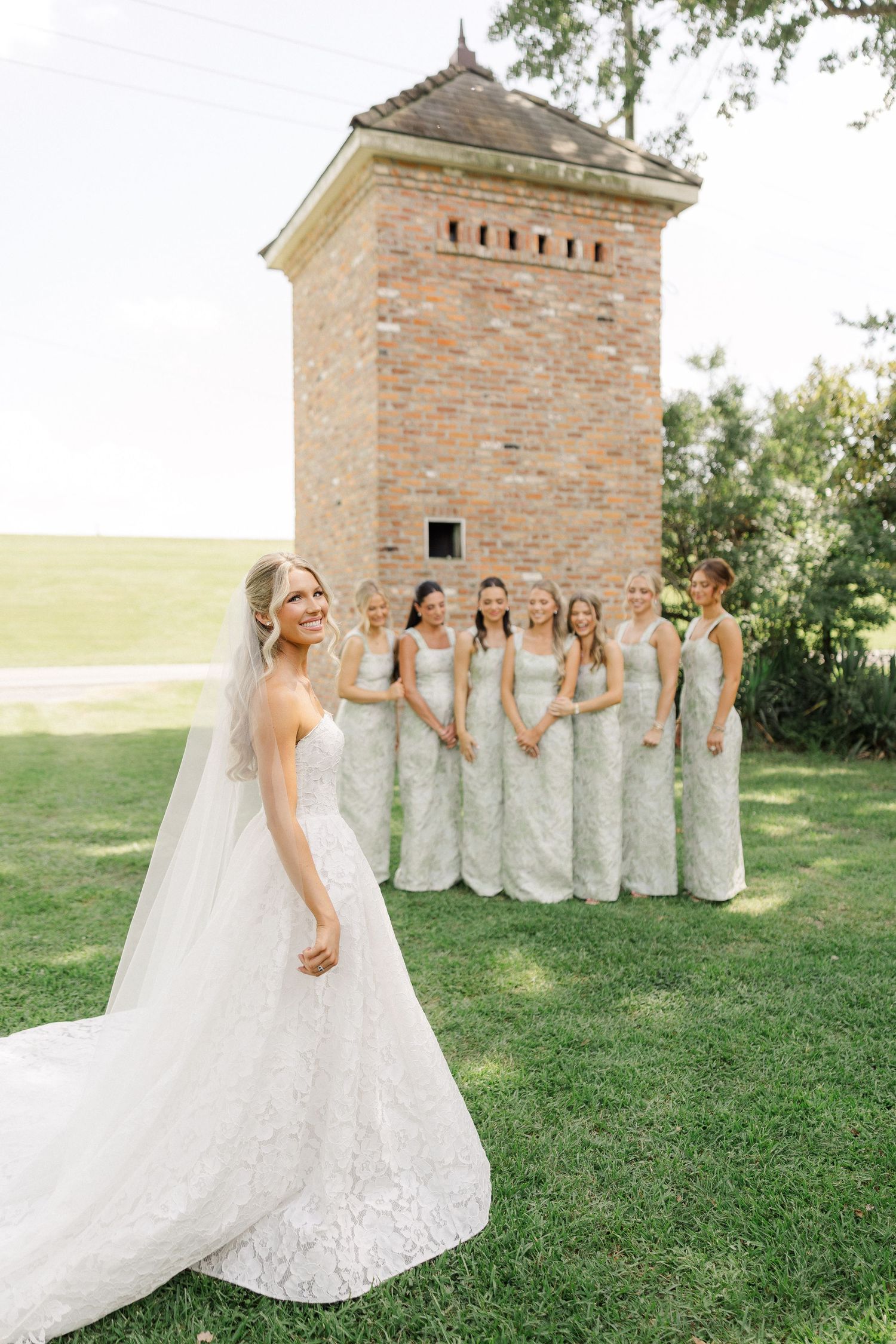 Bride in lace wedding gown with veil poses before bridesmaids in sage green dresses near brick tower.