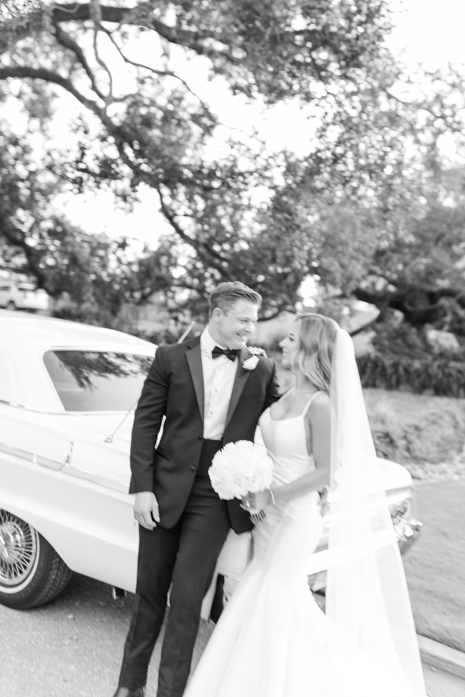 Black and white photo of a bride and groom smiling at each other beside a vintage white car under oak trees.