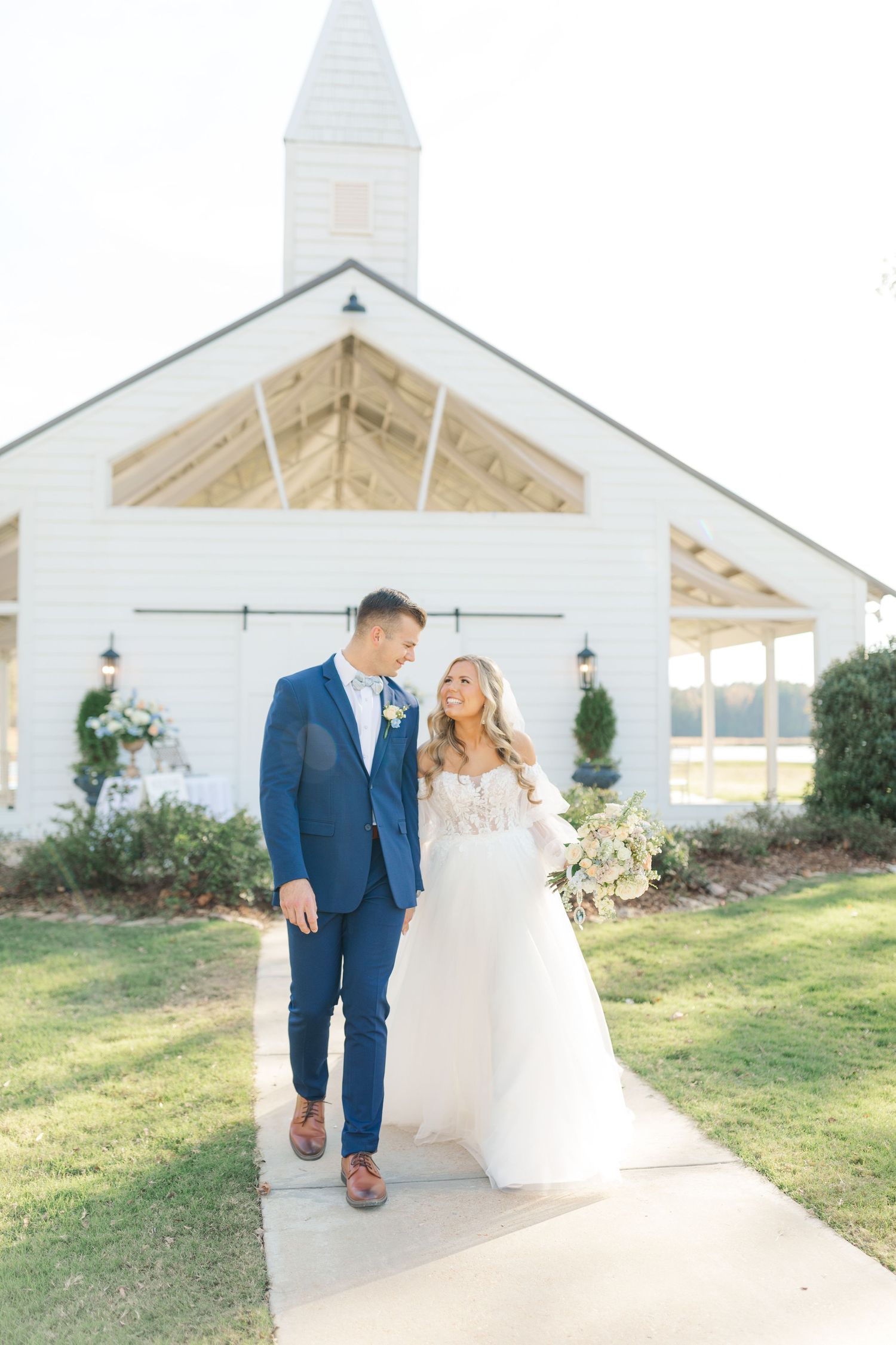 A couple in wedding attire walks along a path in front of a white church with a steeple.