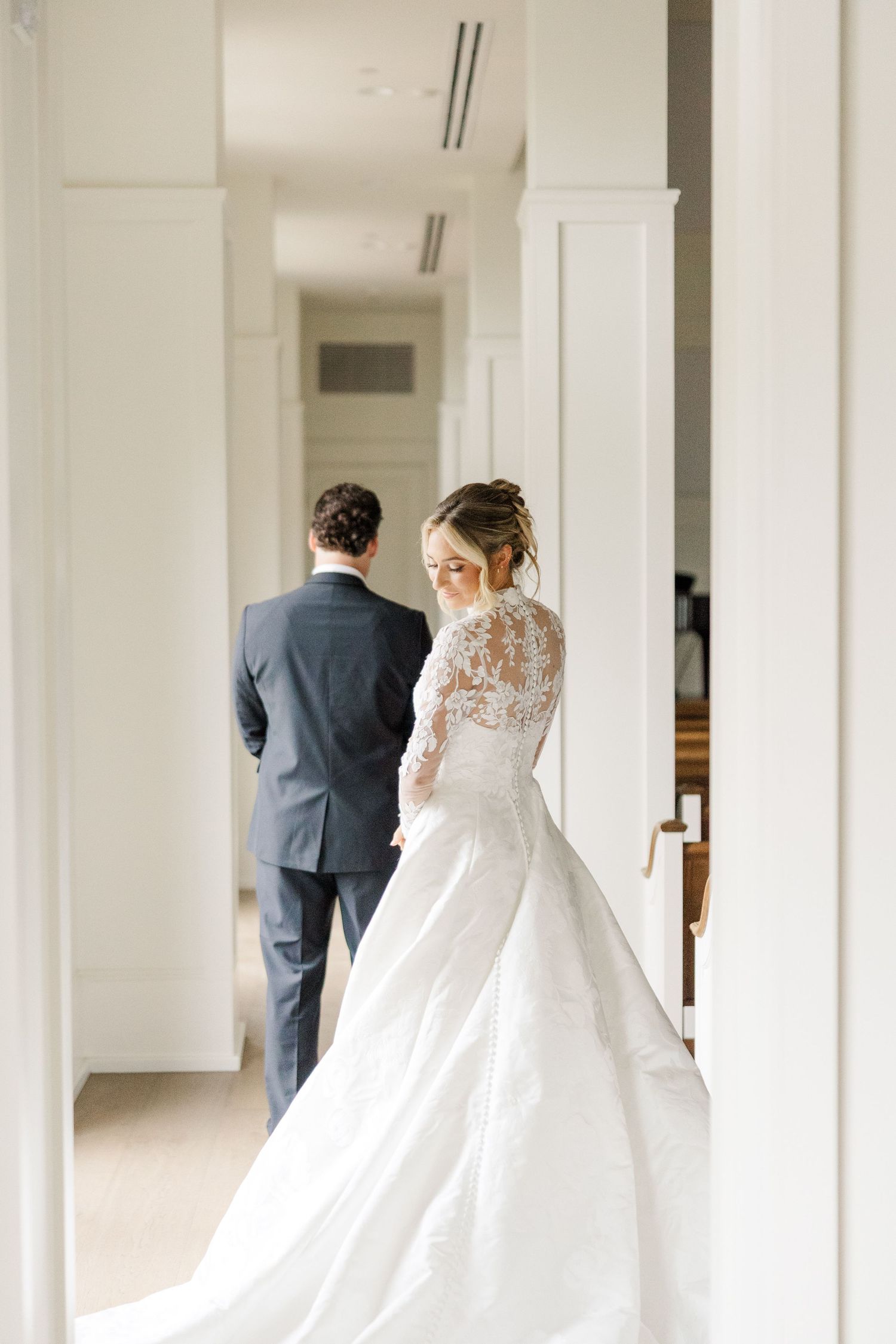 A bride in a flowing white lace gown walks through an elegant white hallway with columns.
