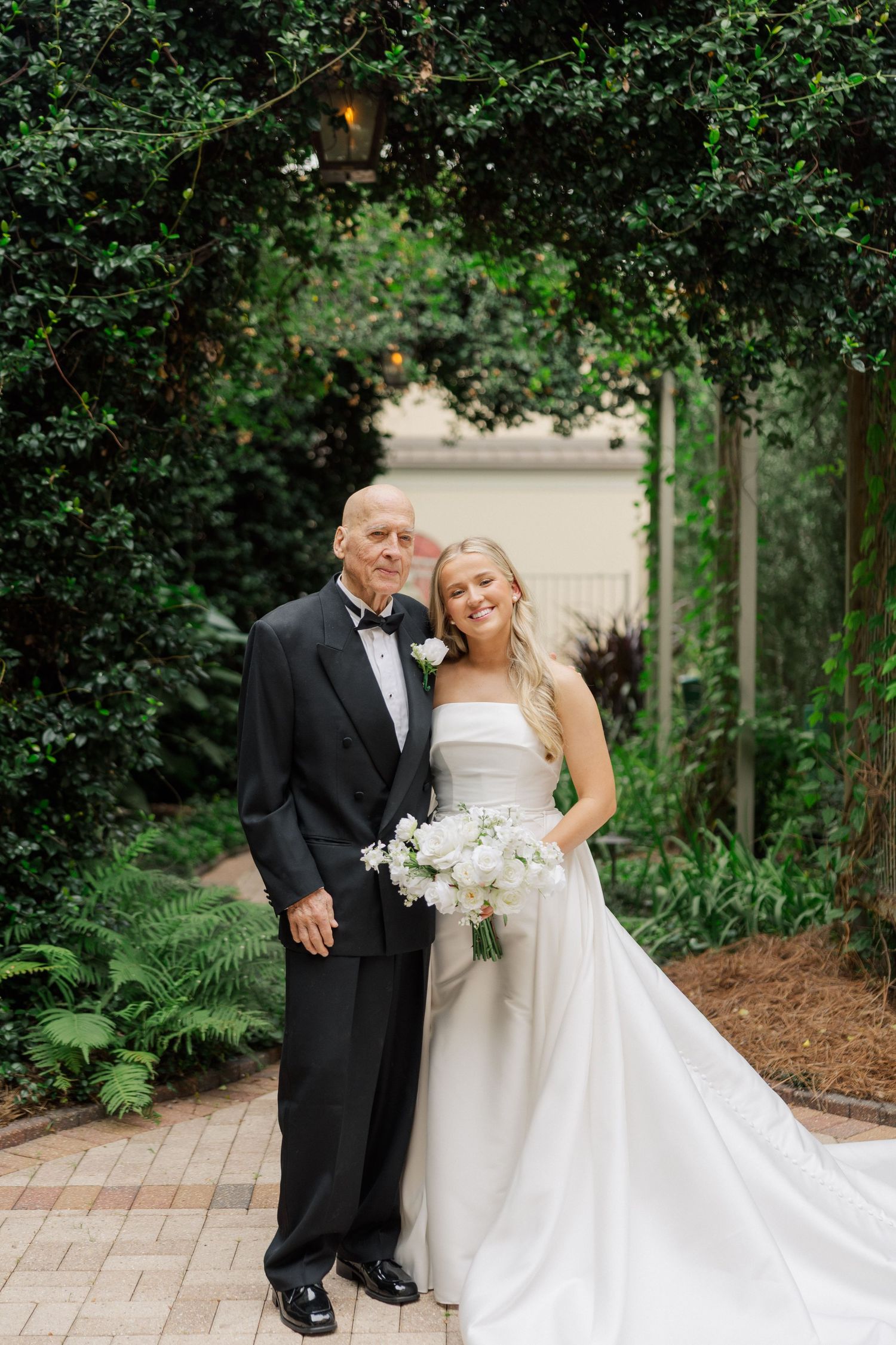 Elegant wedding couple posing together on a garden pathway surrounded by lush greenery.