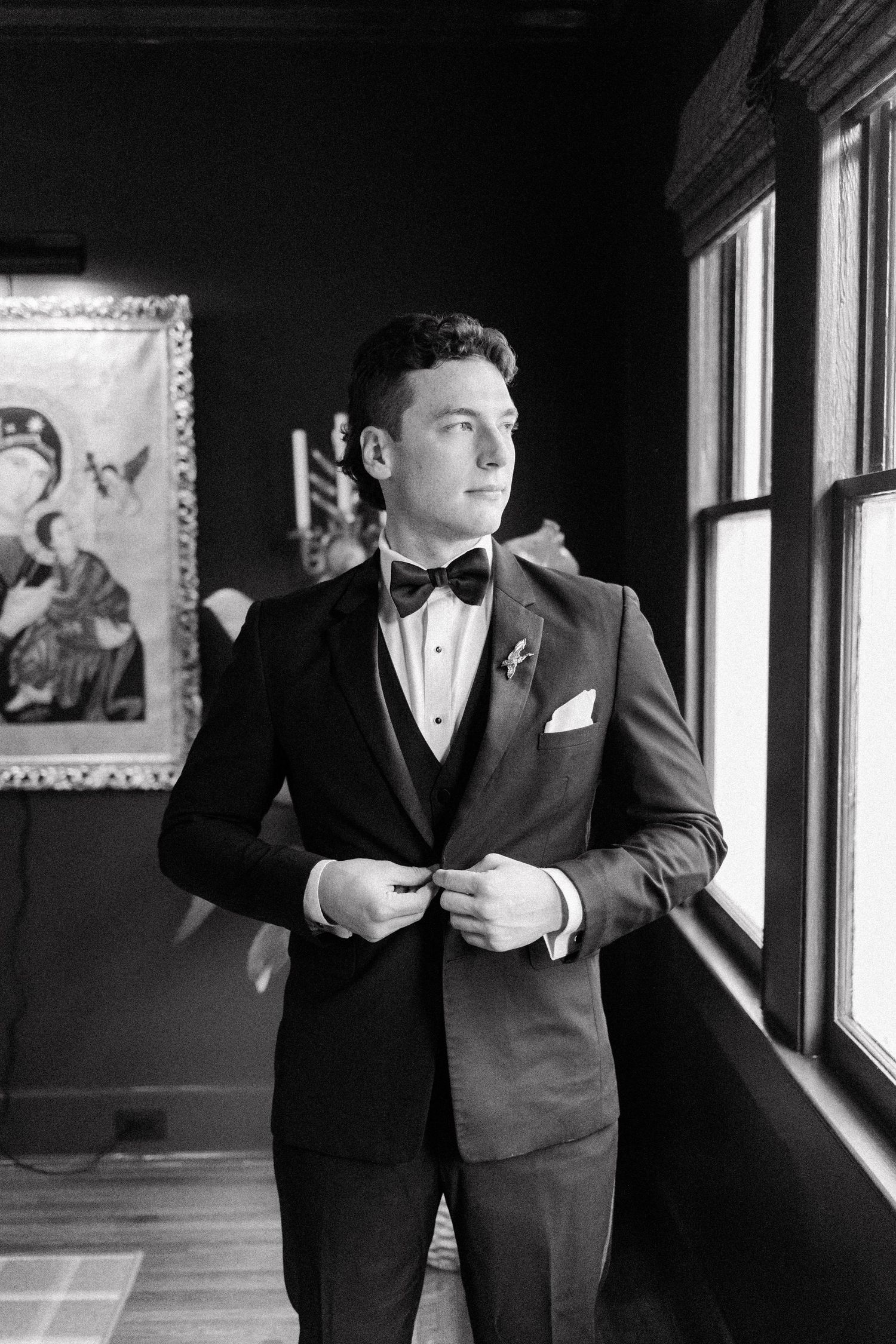 Groomsmen in formal black suits and vests pose by windows in a dark hallway.
