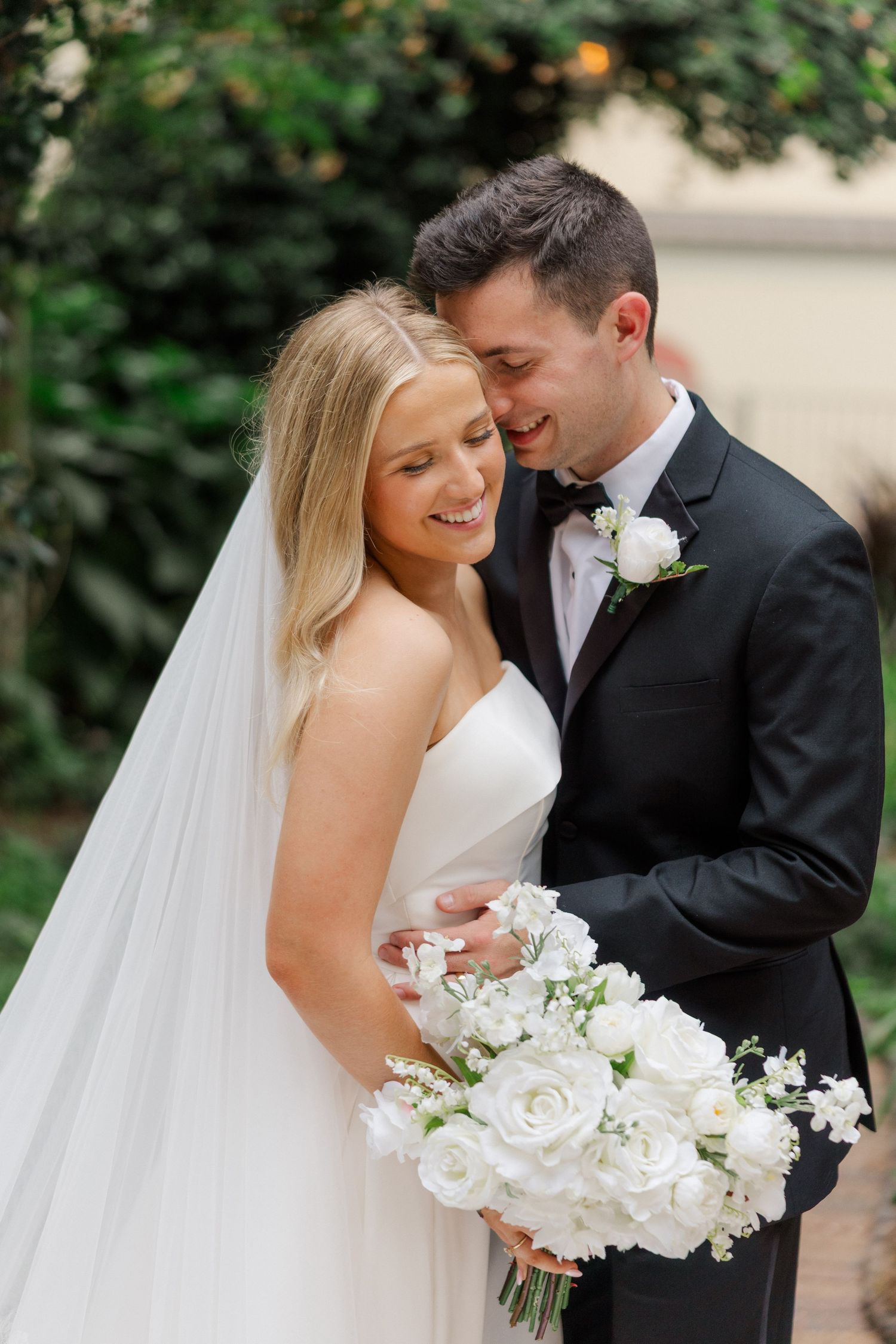 Romantic wedding couple shares intimate moments in garden setting with white floral bouquet and flowing veil.