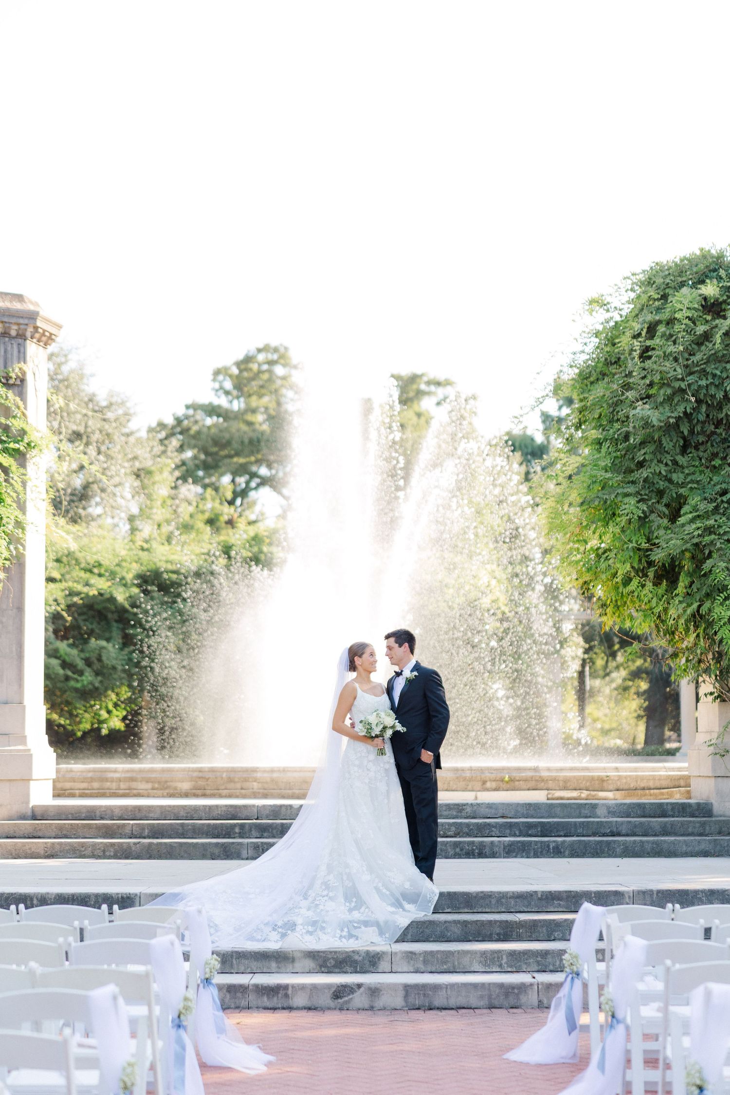 Wedding ceremony setup with white chairs and fountain backdrop in an elegant garden setting.