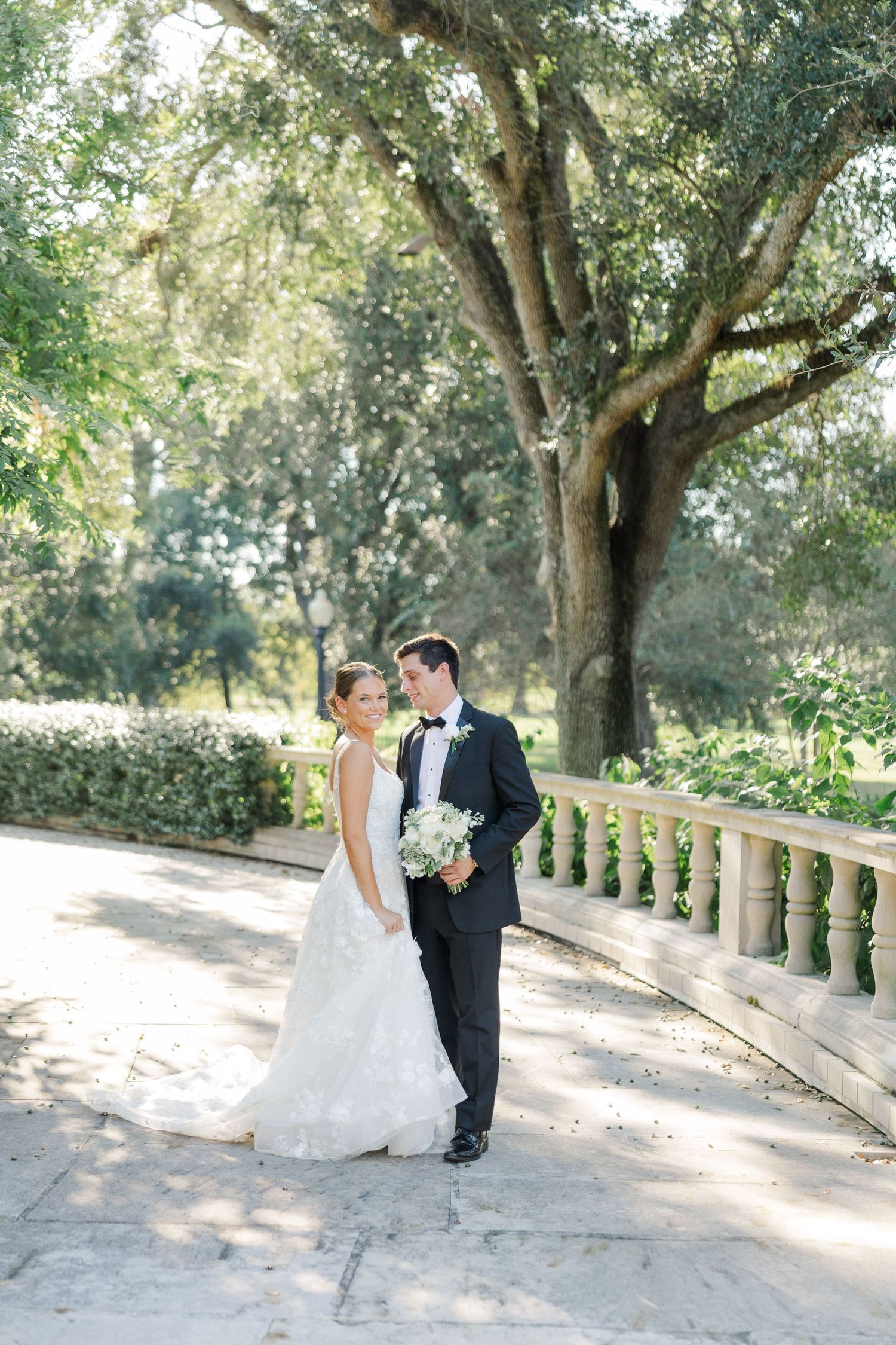 Elegant couple in wedding attire walking along a sunlit stone pathway surrounded by trees.